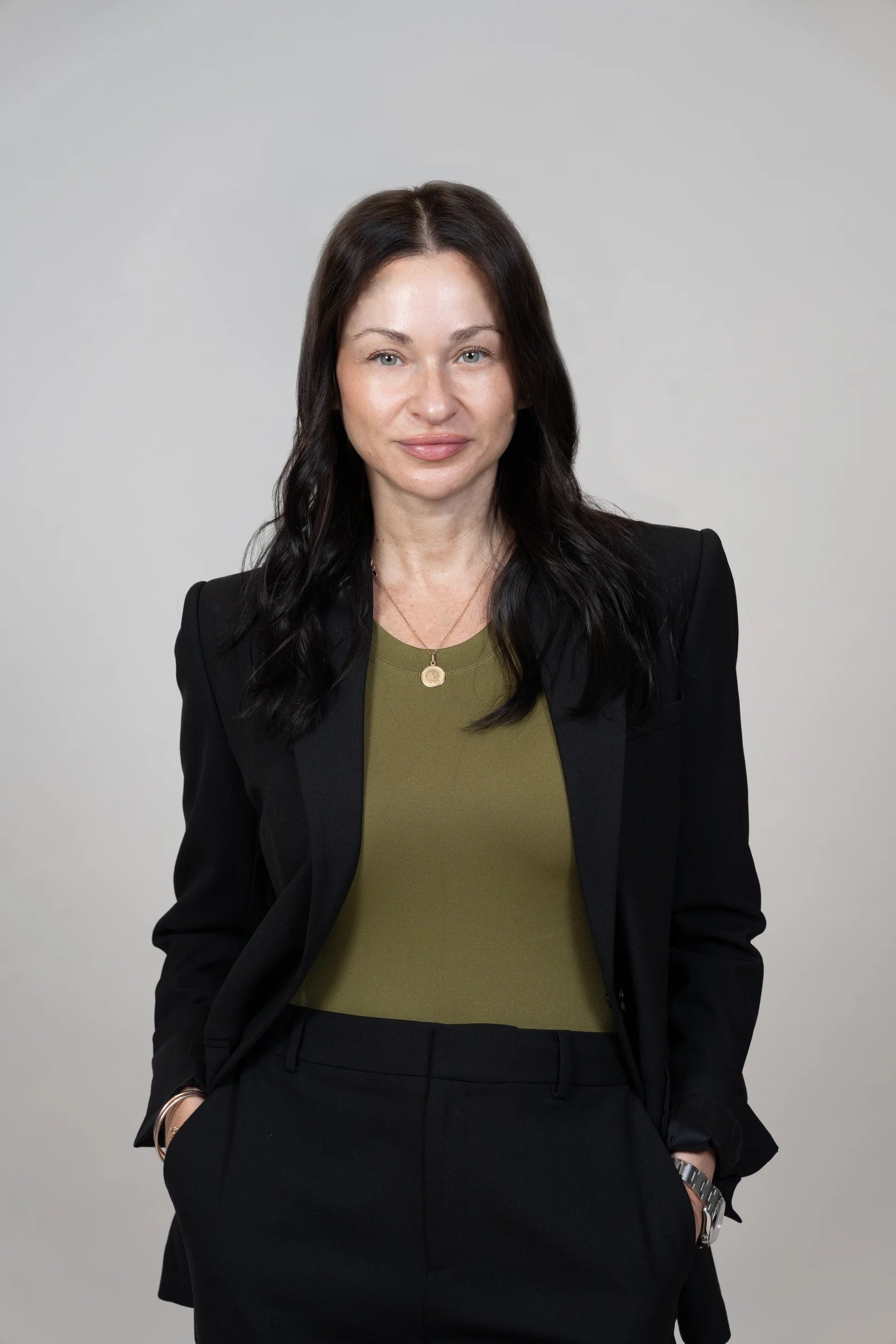 A woman with long dark hair, wearing a black blazer, green top, and black pants, standing against a plain light gray background.
