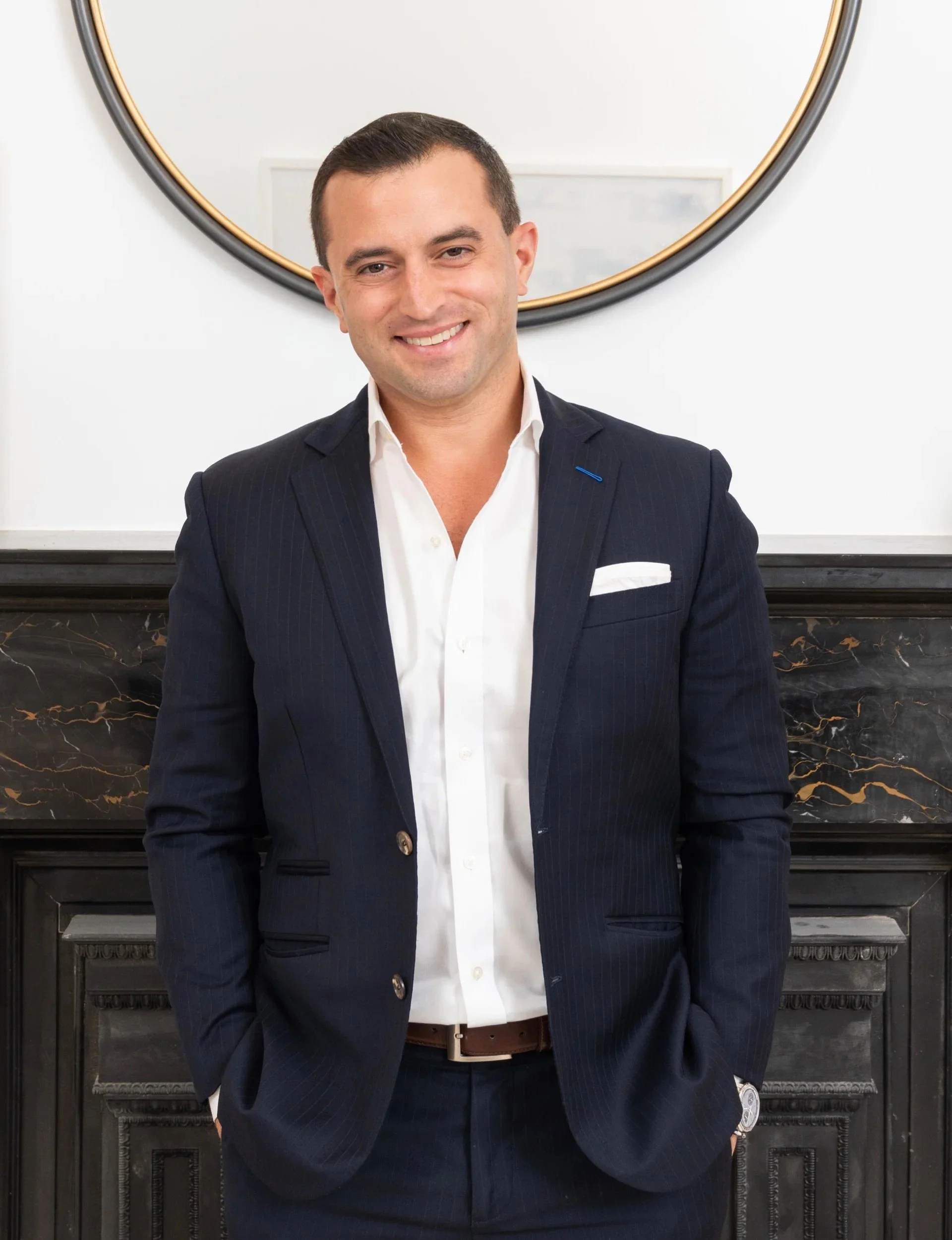 A man in a dark pinstripe suit with a white shirt, standing in front of a black fireplace with marble accents, and a round mirror with a black and gold frame hanging above.