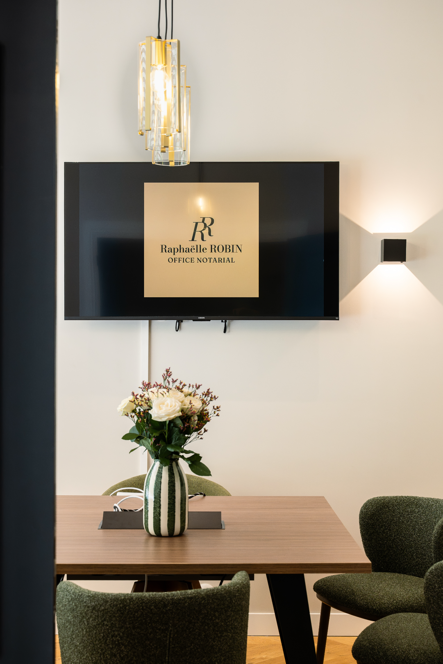 Salle de réunion de l'étude notariale de Raphaëlle ROBIN, avec une télévision affichant le nom de l'office notarial, une table en bois, des chaises vertes, une lampe suspendue élégante et un vase avec des fleurs sur la table.