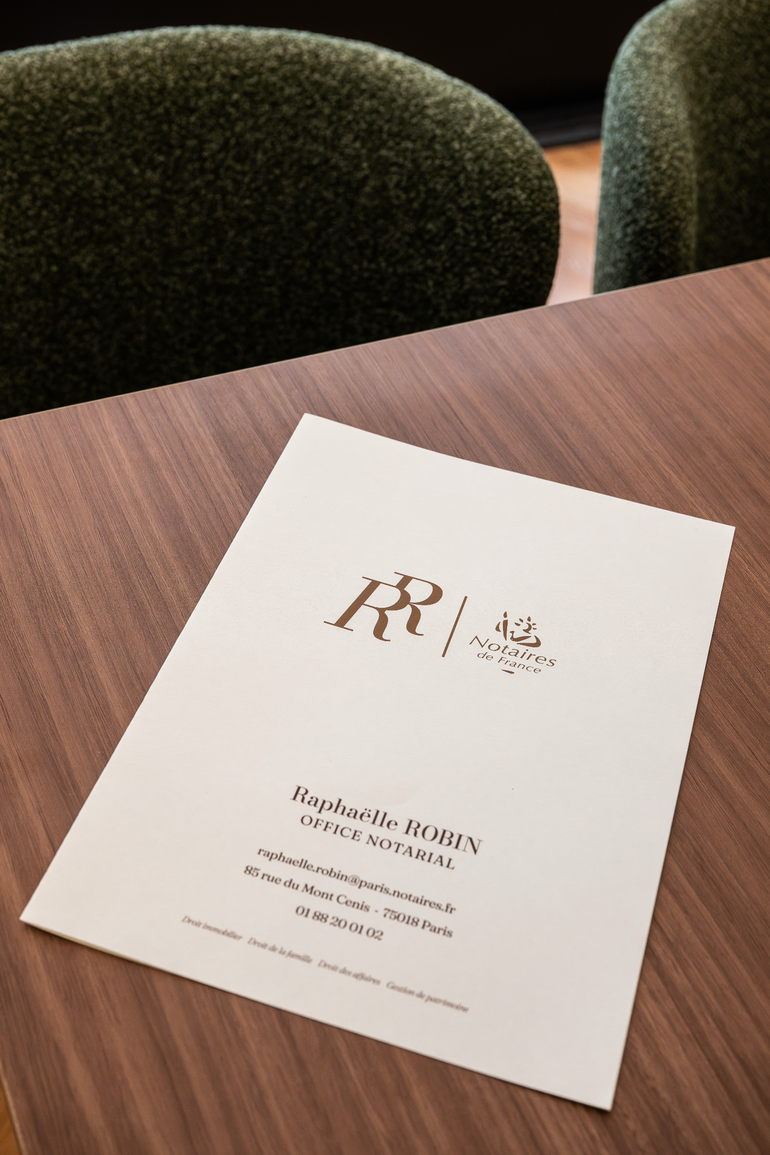 Carte de visite de la notaire Raphaëlle ROBIN, ses coordonnées et l'adresse de son étude à Paris 18, posée sur un bureau en bois avec deux chaises vert foncé en arrière-plan.