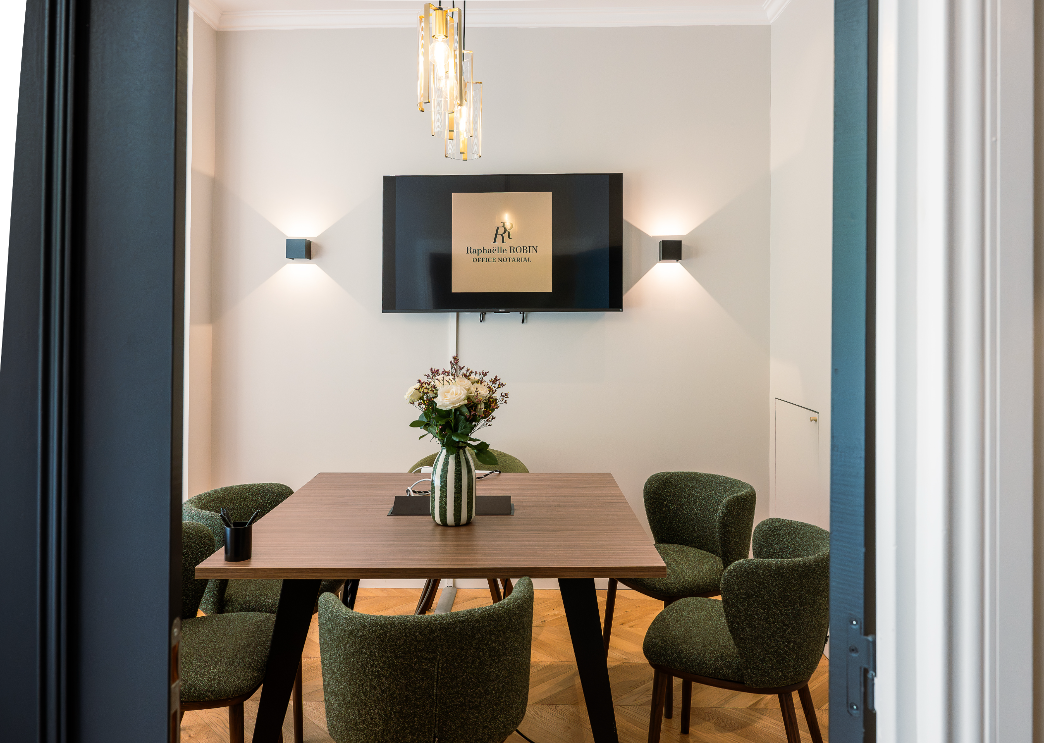 Salle de réunion de l'office notarial de Raphaëlle ROBIN, table en bois avec une vase contenant des fleurs, murs blancs, éclairage moderne avec lampes murales et suspension.