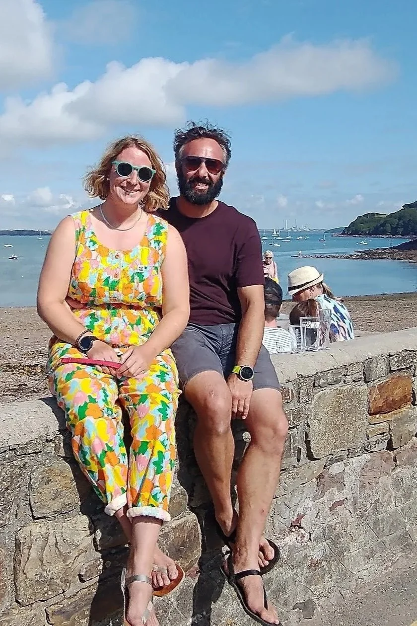 A woman and a man sitting on a stone wall by the beach, smiling, with boats in the water and a cloudy sky in the background.