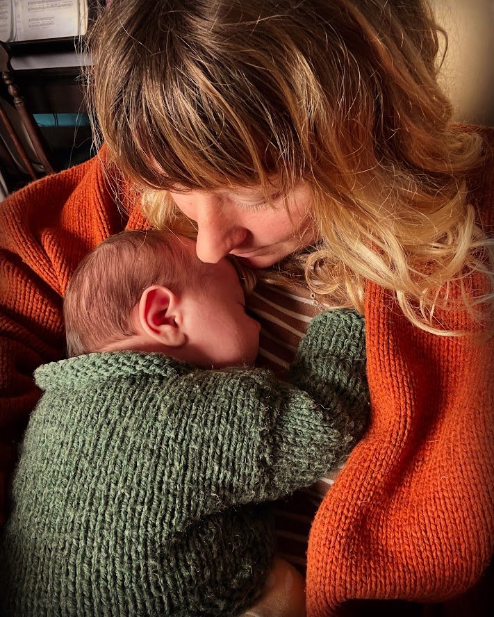 A woman with wavy blonde hair close to a sleeping baby wearing a green sweater, both resting their foreheads together in an intimate moment.