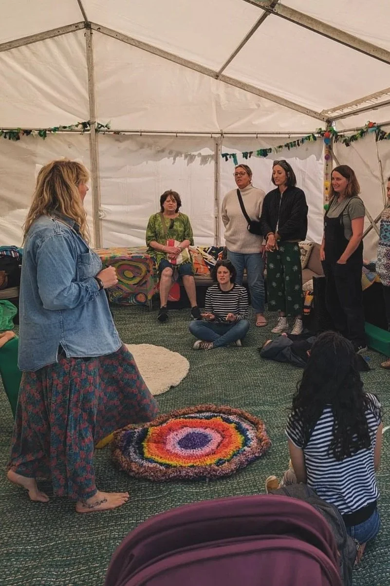 A woman leading a group of people in song inside a white tent with colourful decorations. Some are sitting on the floor while others stand, listening attentively.