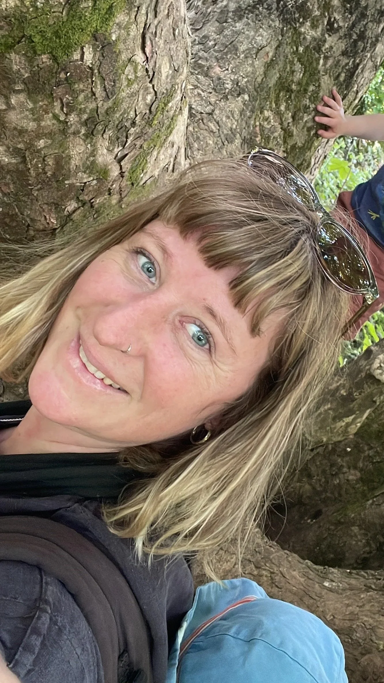 A woman with blonde hair and a nose piercing smiling for a photo outdoors, leaning against a tree with moss, with a child’s hand on the tree in the background.