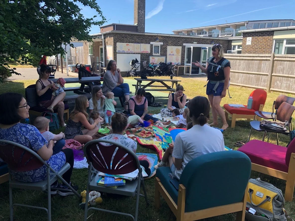 A group of adults and children gathered outdoors in a backyard, sitting on blankets and chairs, listening to a woman standing and speaking, with playground equipment and houses in the background.
