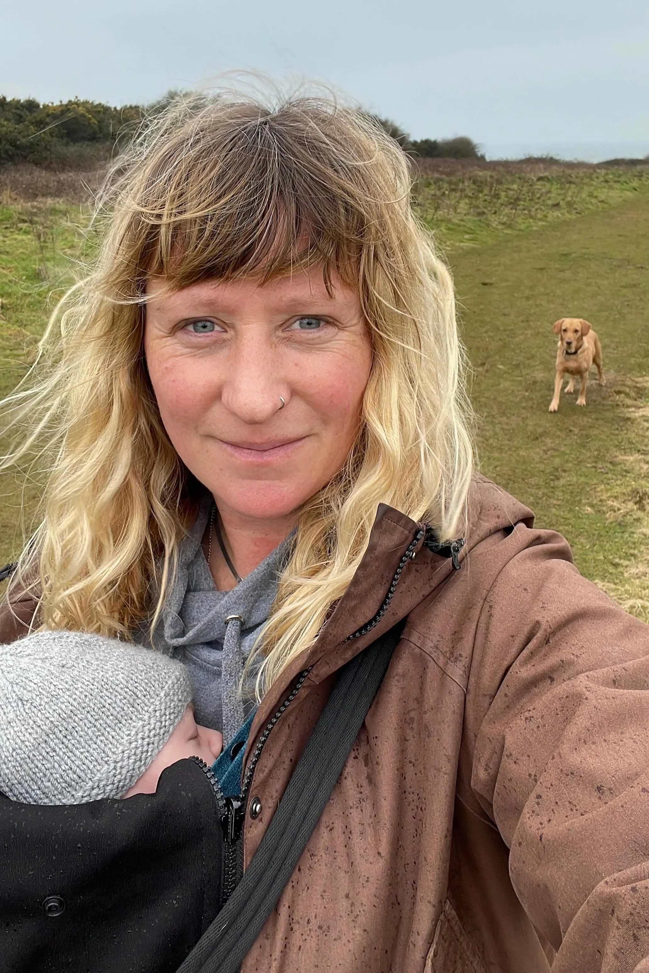 A woman with blonde hair and blue eyes taking a selfie outdoors with a dog in the background on a grassy path. The woman is wearing a brown jacket and a gray hoodie underneath, and the dog appears to be a Labrador Retriever.