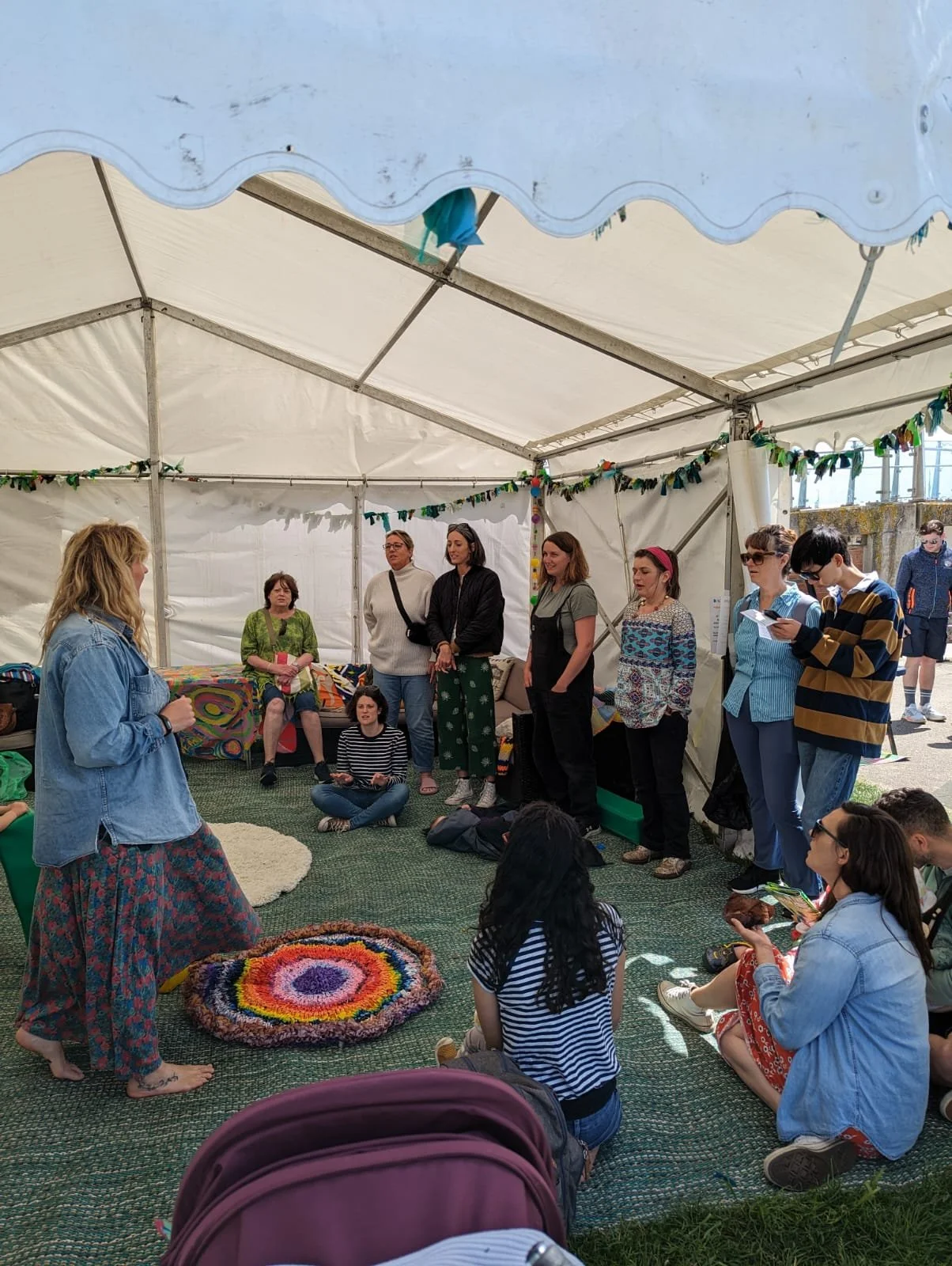 A woman leading a group of people in song inside a white tent with colourful decorations. Some are sitting on the floor while others stand, listening attentively.