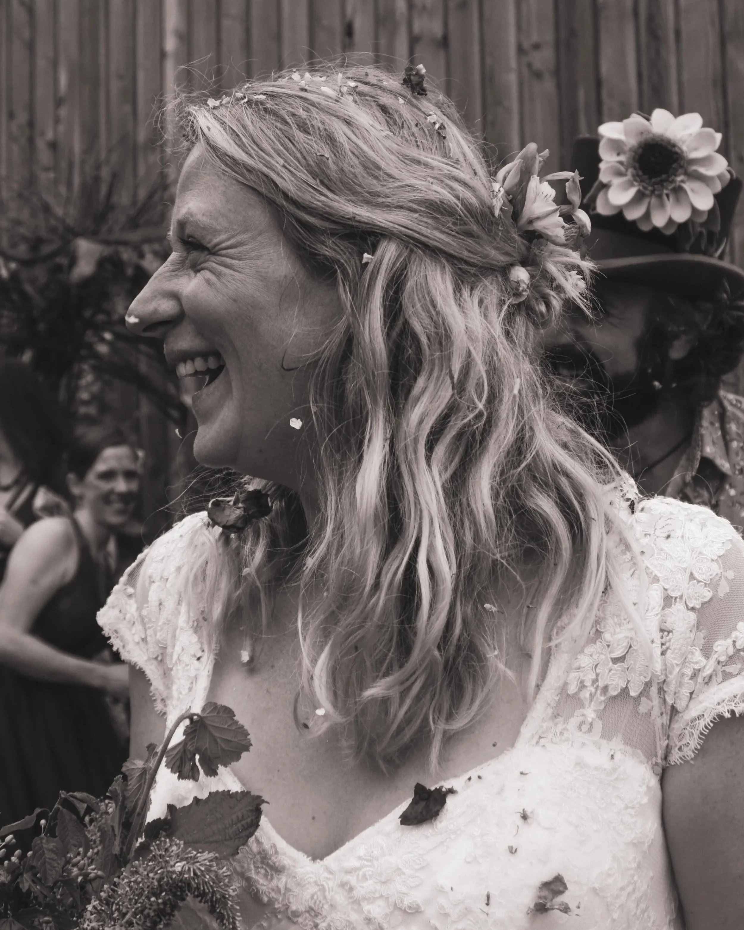 A woman with long wavy hair, wearing a lace dress and a floral crown, smiling as she poses outdoors with a wooden fence in the background. There are flowers and leaves in the foreground.