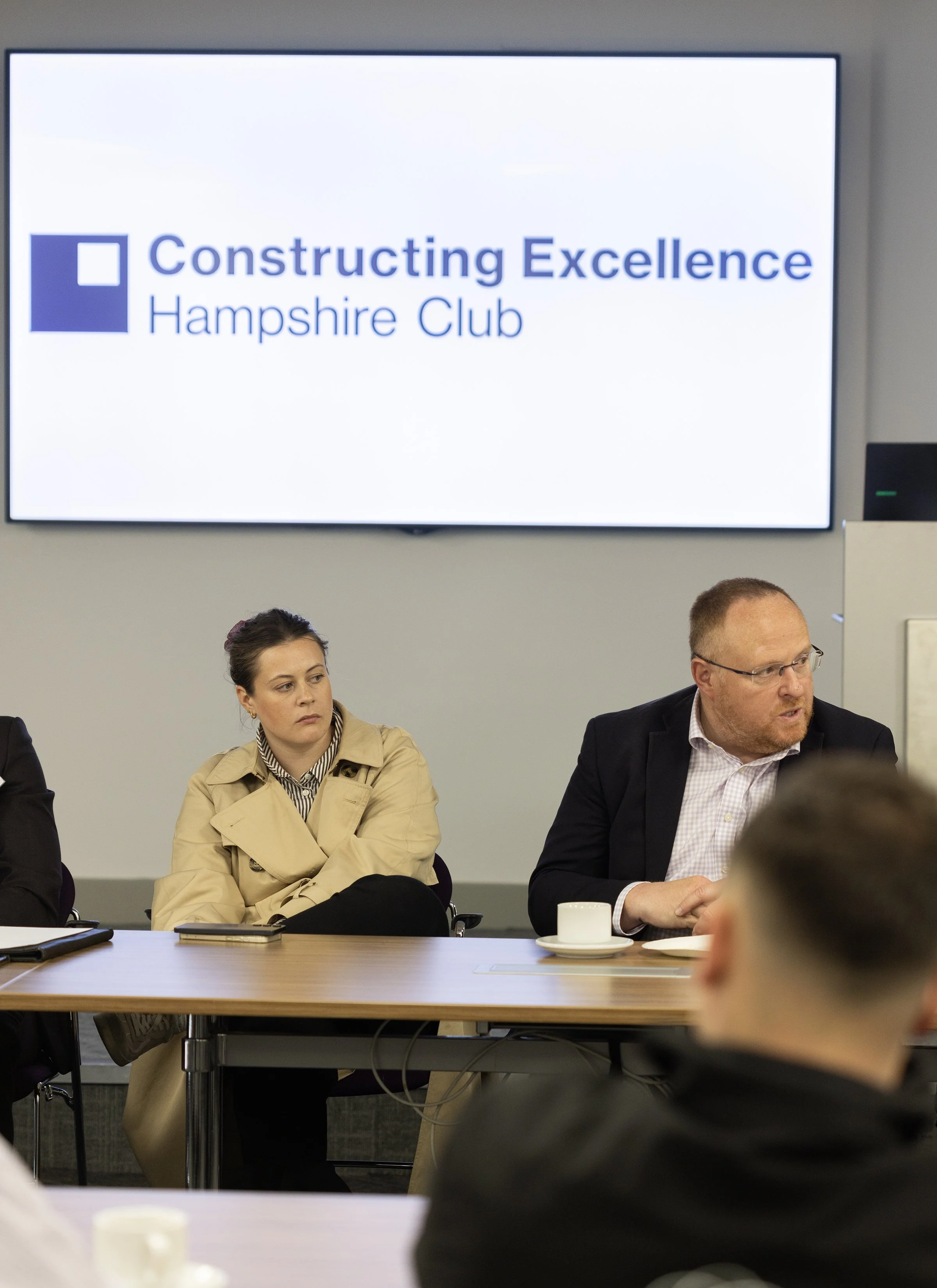 A group of people sitting at a conference table during a meeting, with a large screen in the background displaying a logo and text that reads 'Constructing Excellence Hampshire Club.'