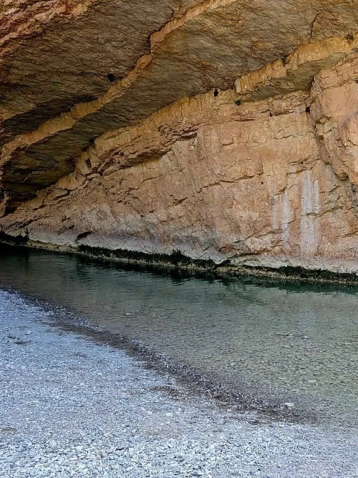 Une grotte ou falaise rocheuse avec un rivage de galets, où l'eau calme touche la roche.