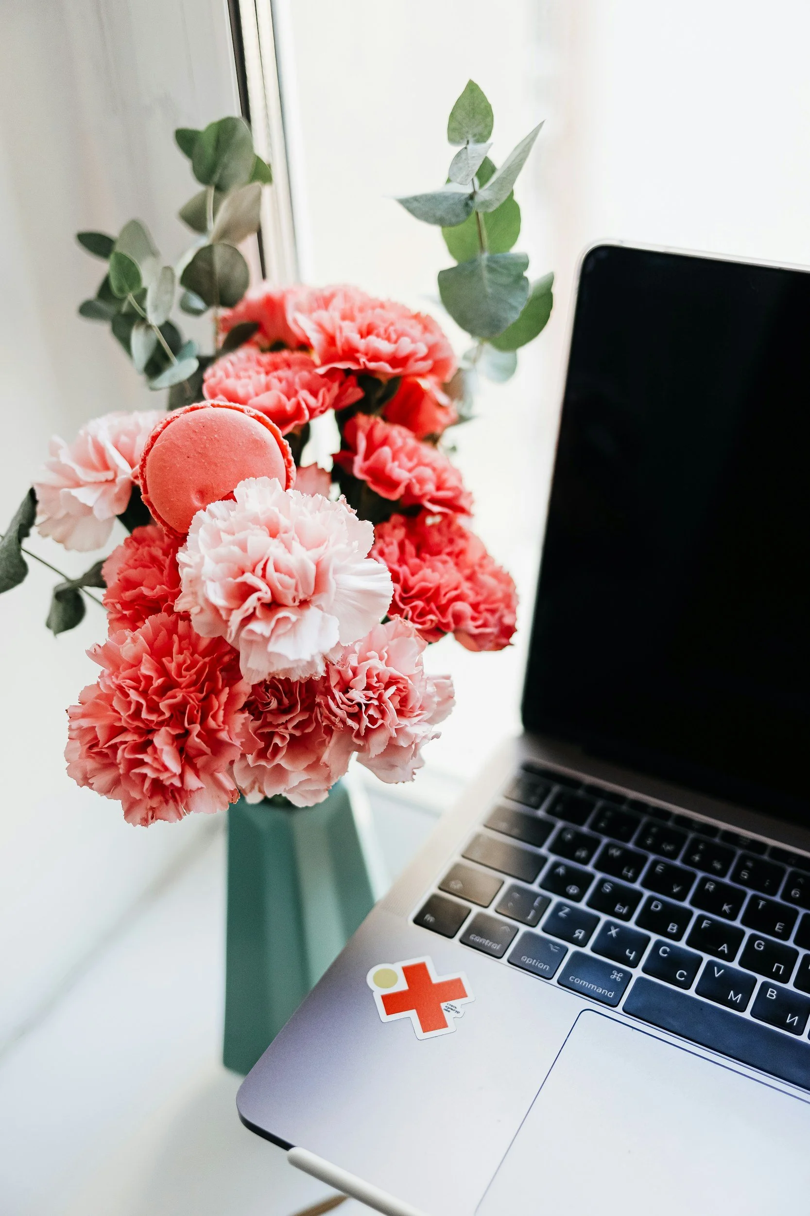 Close-up of a laptop with a sticker, next to a bouquet of pink carnations and eucalyptus leaves on a white table near a window.