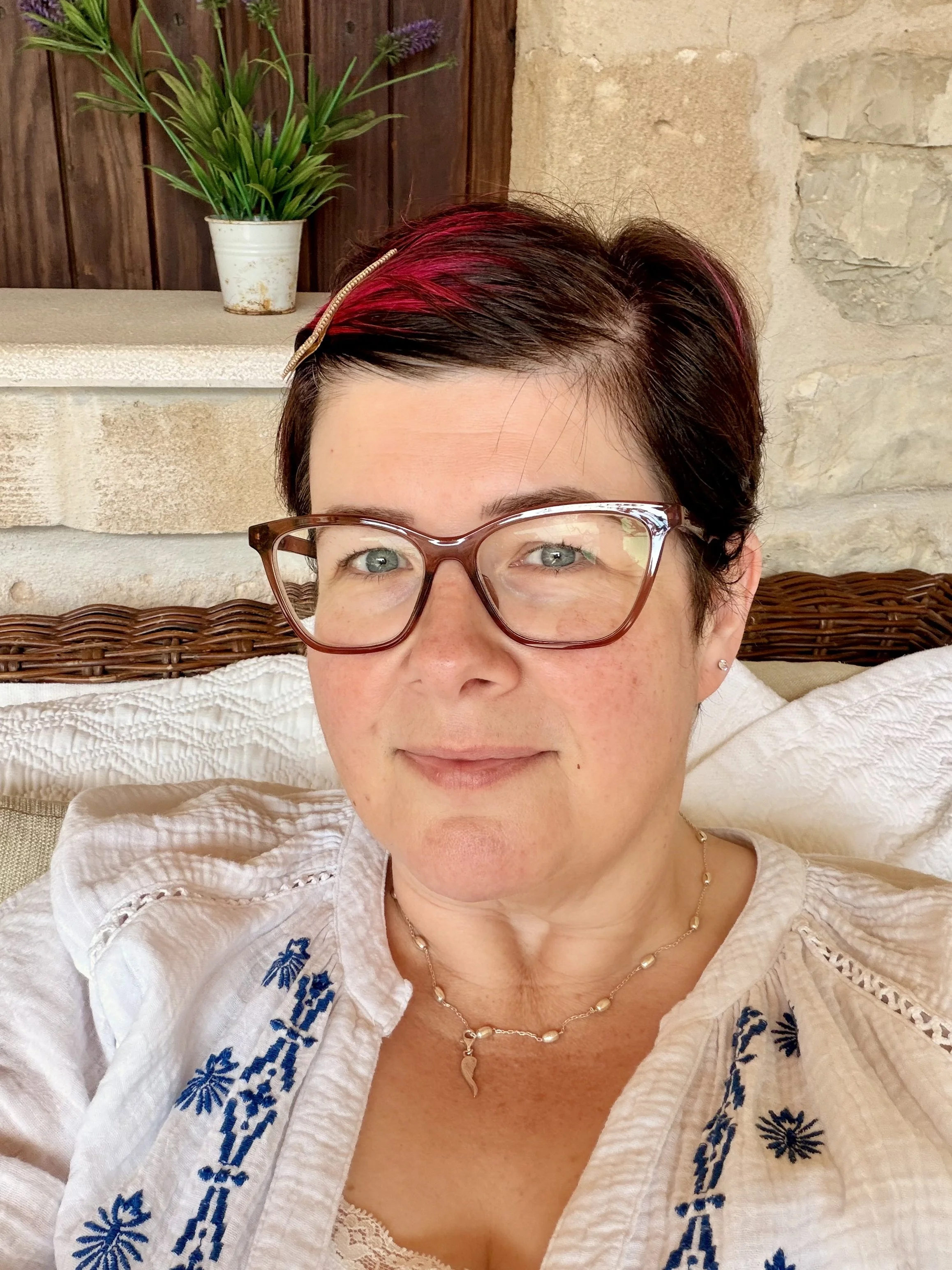 A woman with short dark hair with pink highlights, wearing glasses and a white embroidered blouse, sitting in a cozy setting with a stone and wood background, and a potted plant and decorative items behind her.