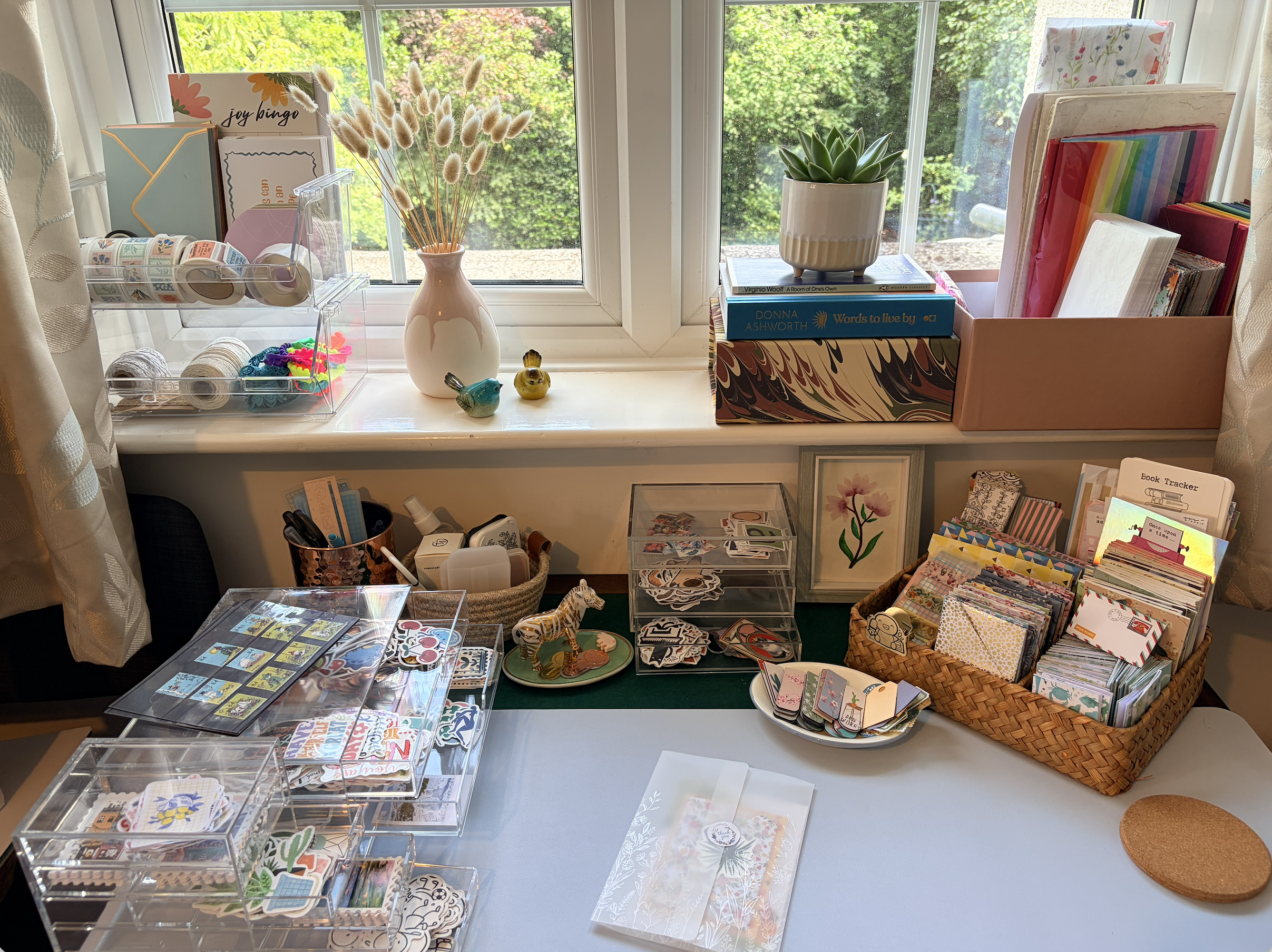 A collection of stationery items, notebooks, stickers, and craft supplies displayed on a white table with a window showing green trees outside. Decorative objects like a vase with dried flowers and small ceramic figurines are also visible.