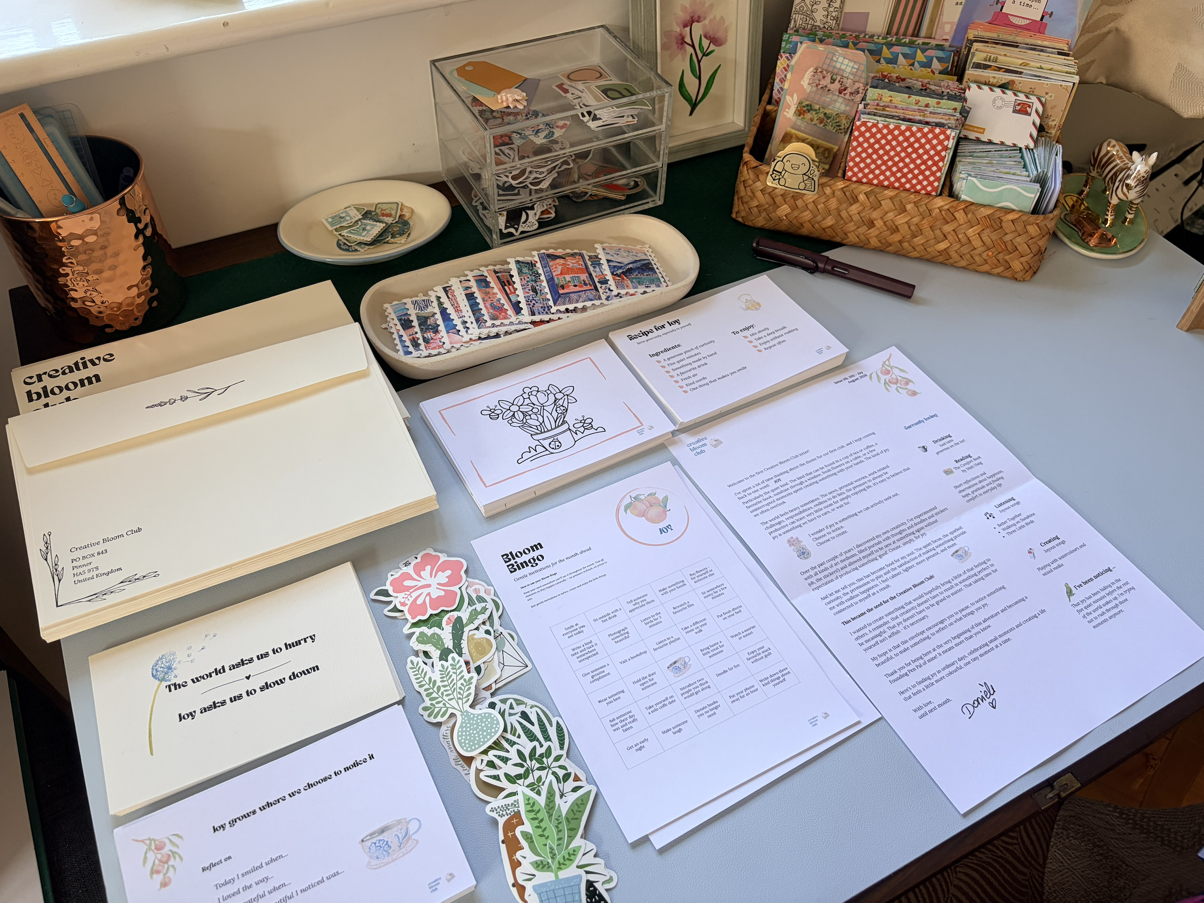 A desk decorated with various papers, stickers, and craft supplies, including a basket of cards, a tray of colorful postage stamps, and a stack of printed documents, with decorative plants and stationery items surrounding them.