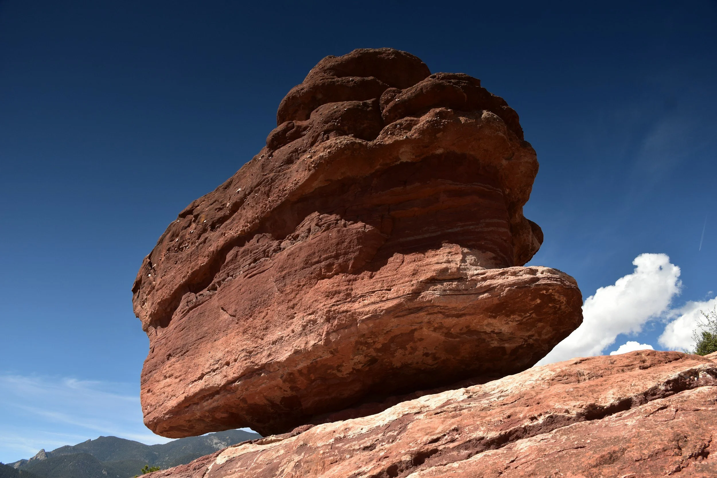 A large, reddish-brown rock formation with stacked layers, under a clear blue sky with some clouds and distant mountains visible in the background.