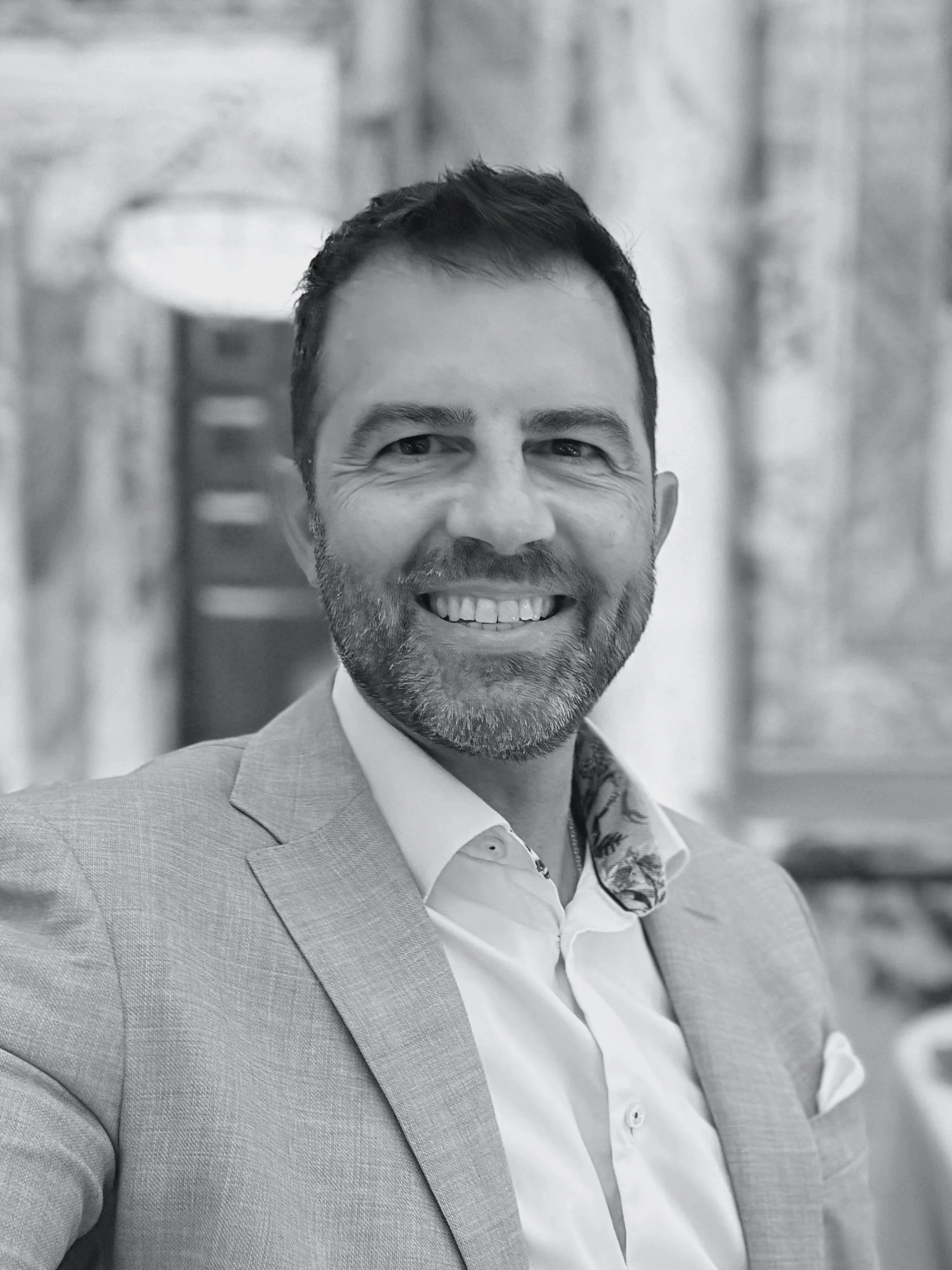 Close-up of a man with short dark hair and a beard, smiling, wearing a light-colored suit jacket, white shirt, and patterned scarf, in an indoor setting.