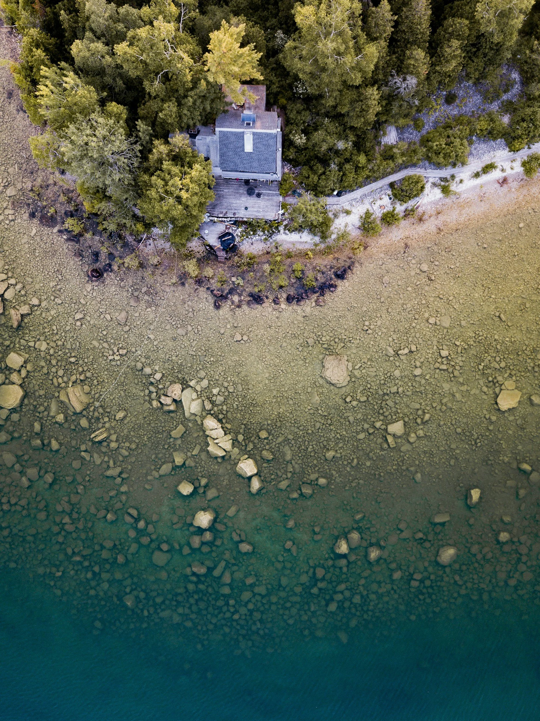 An aerial view of a house surrounded by dense trees with a wooden deck, located along a body of water with rocks and pebbles along the shoreline.