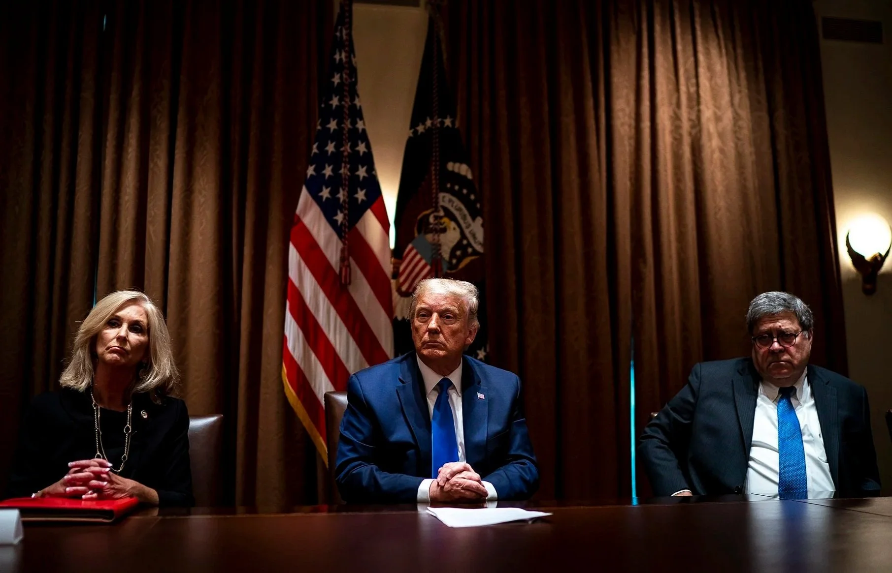 Three individuals, a woman on the left, Donald Trump in the center, and a man on the right, sitting at a conference table in a room with brown curtains and two American flags behind them.
