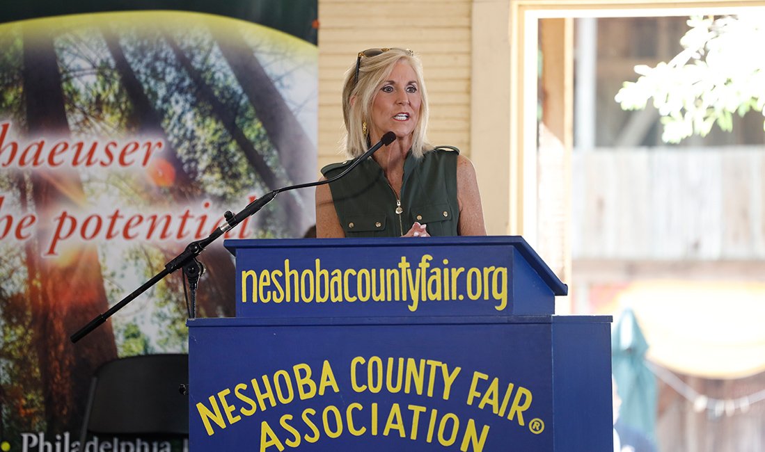 A woman speaking at a podium with a banner in the background and a sign that reads 'neshobacountyfair.org' and 'Neshoba County Fair Association'.