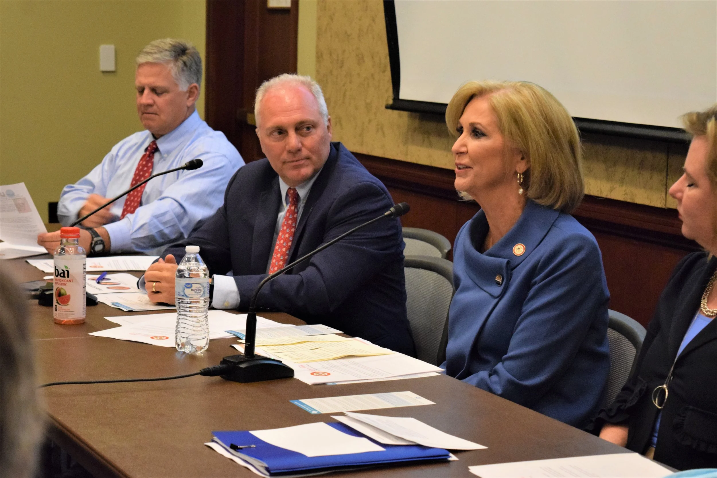 A group of four people sitting at a conference table during a meeting, with documents, water bottles, and a microphone in front of them, and a blank white screen behind them.