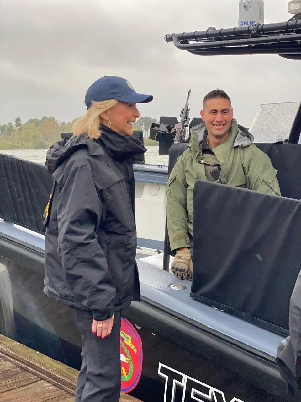 A woman and a man on a boat, smiling and talking. The woman is wearing a navy cap and rain jacket, and the man is in a green military-style jacket. The boat has patrol markings and belongs to the Texas Department of Public Safety, as indicated by the logo on the side.