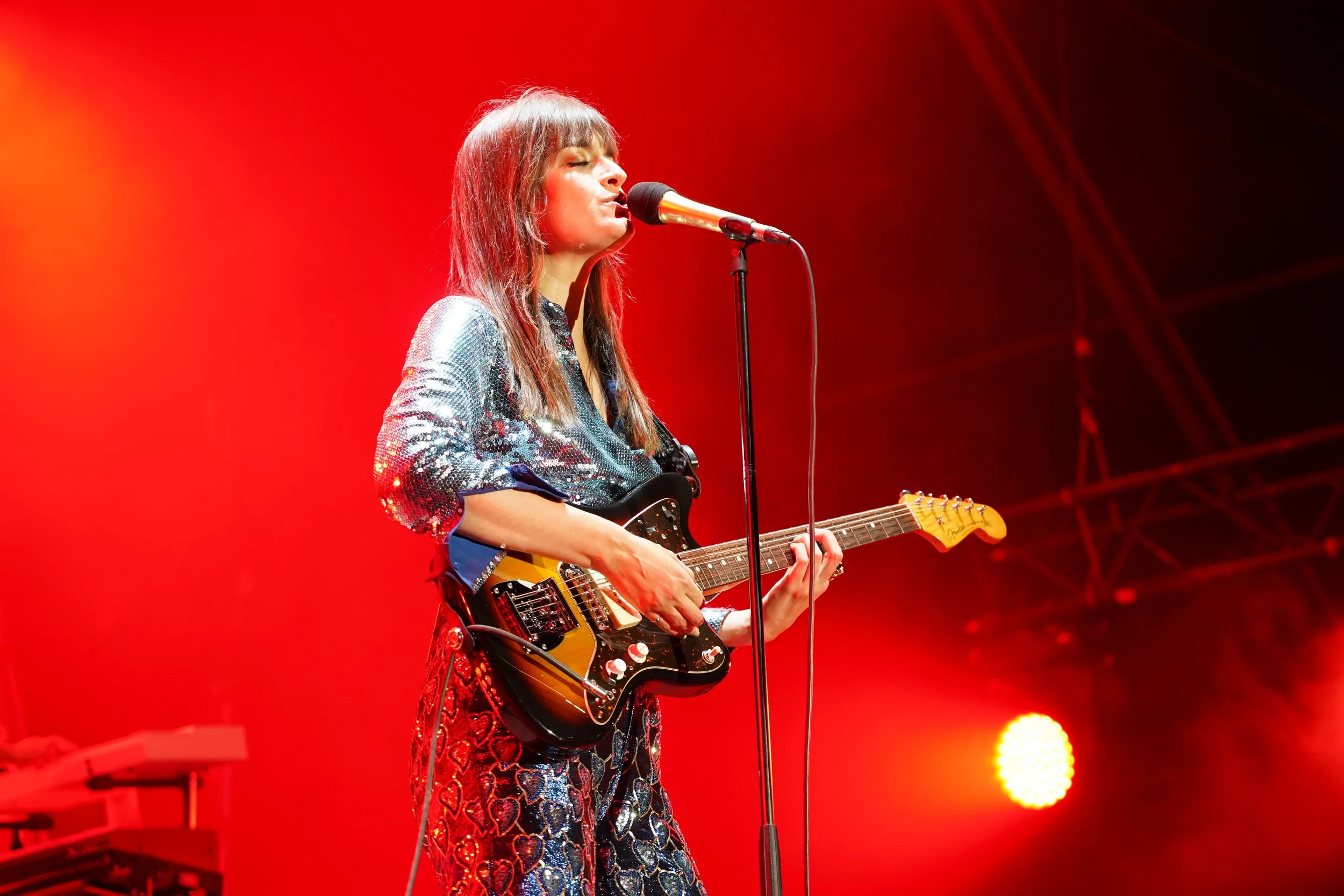 Une femme joue de la guitare électrique lors d'un concert, avec un fond rouge et des lumières chaudes.