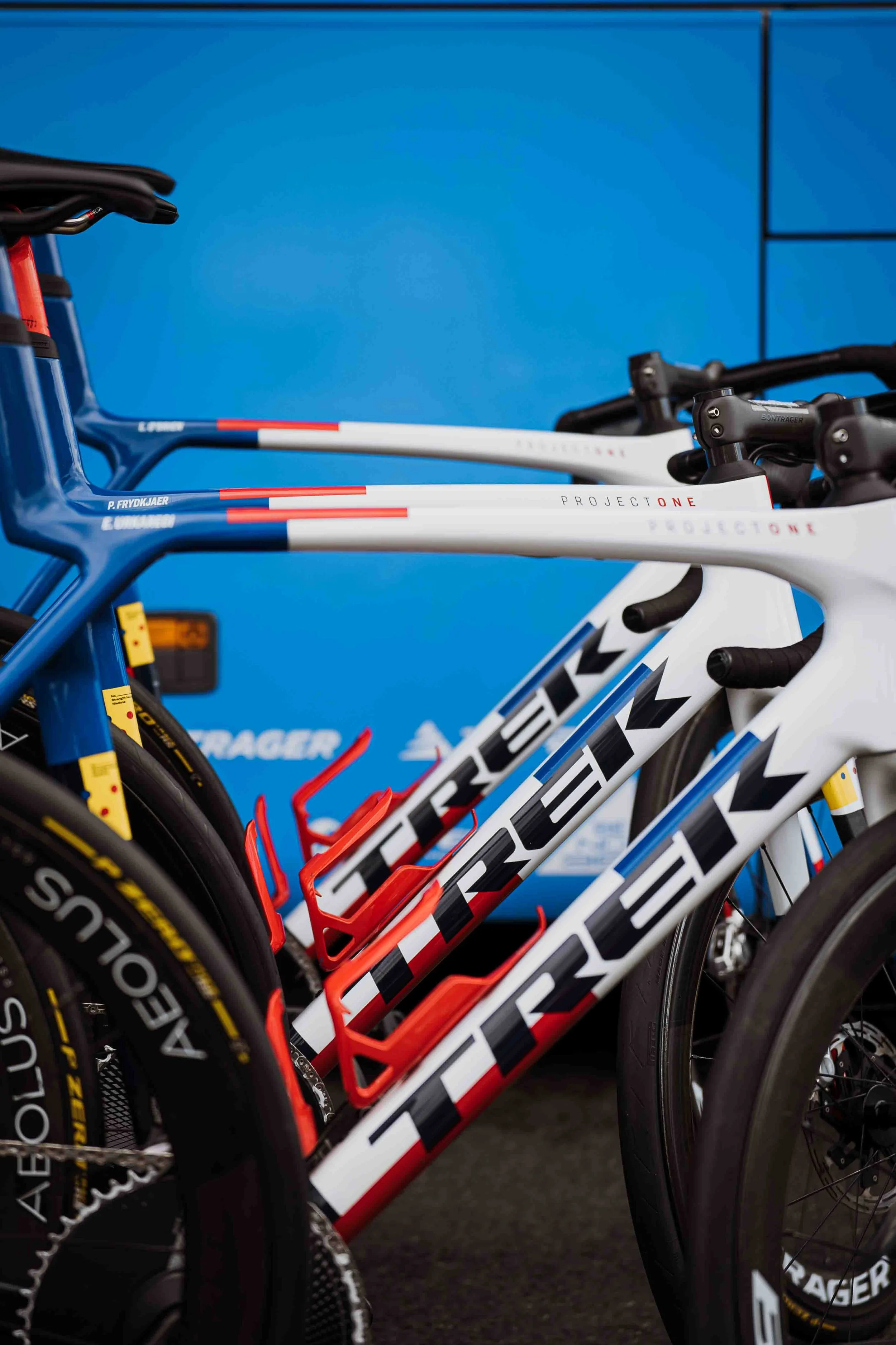 Des vélos de course alignés côte à côte, avec des cadres principalement blancs, rouges et bleus, dans un espace intérieur avec un mur bleu en arrière-plan.