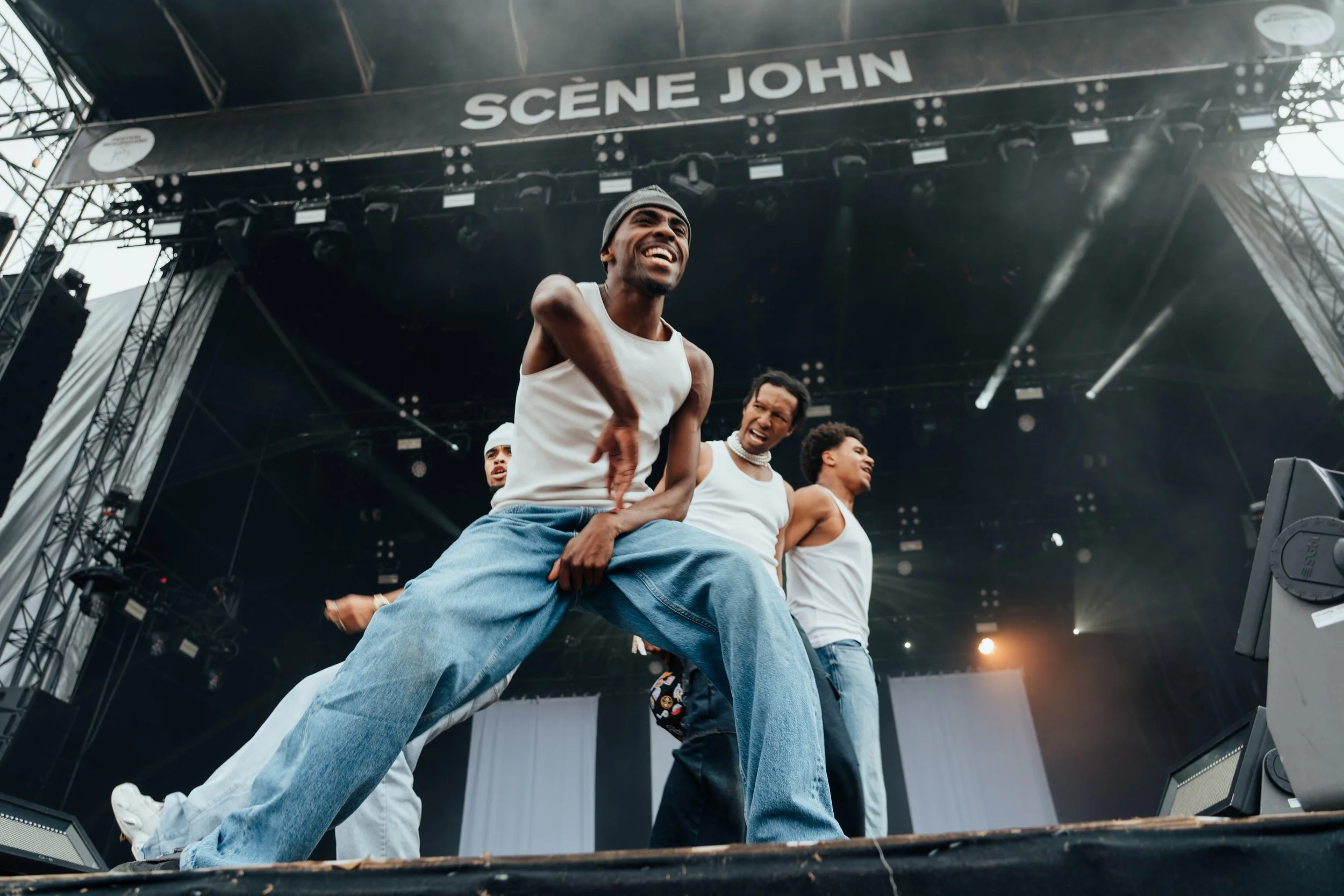 Groupe de personnes dansant sur scène lors d'un spectacle en plein air, avec un panneau 'SCENE JOHN' en arrière-plan.