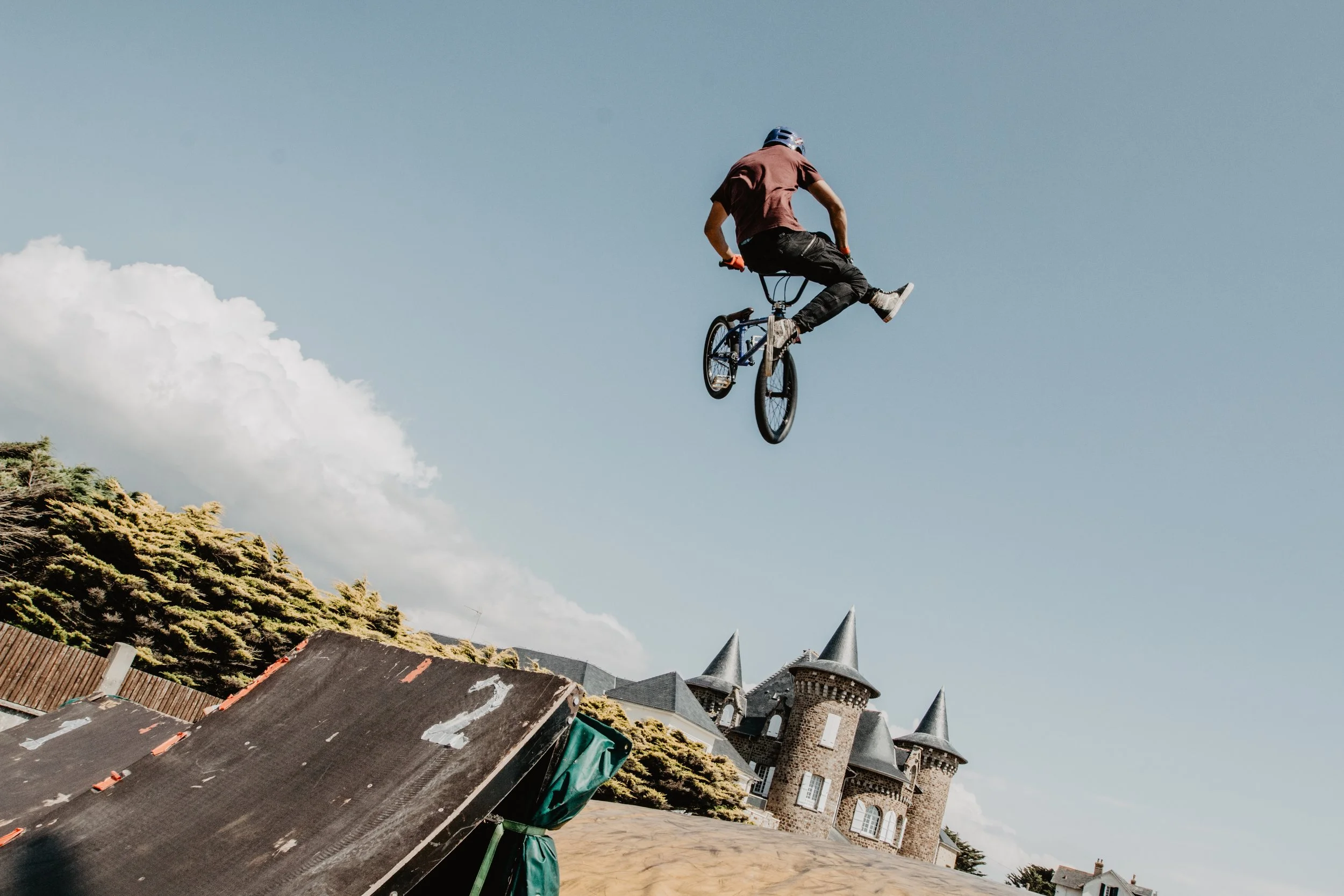 Un jeune homme fait du BMX en sautant dans le ciel près d'un château avec des tours en pierre et des toits pointus, sous un ciel bleu avec quelques nuages.