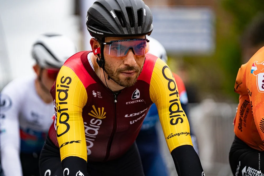 Un cycliste portant un casque noir, des lunettes de soleil roses, et un maillot rouge et jaune avec les mots 'Sop 탈' sur les manches, semble être en course.