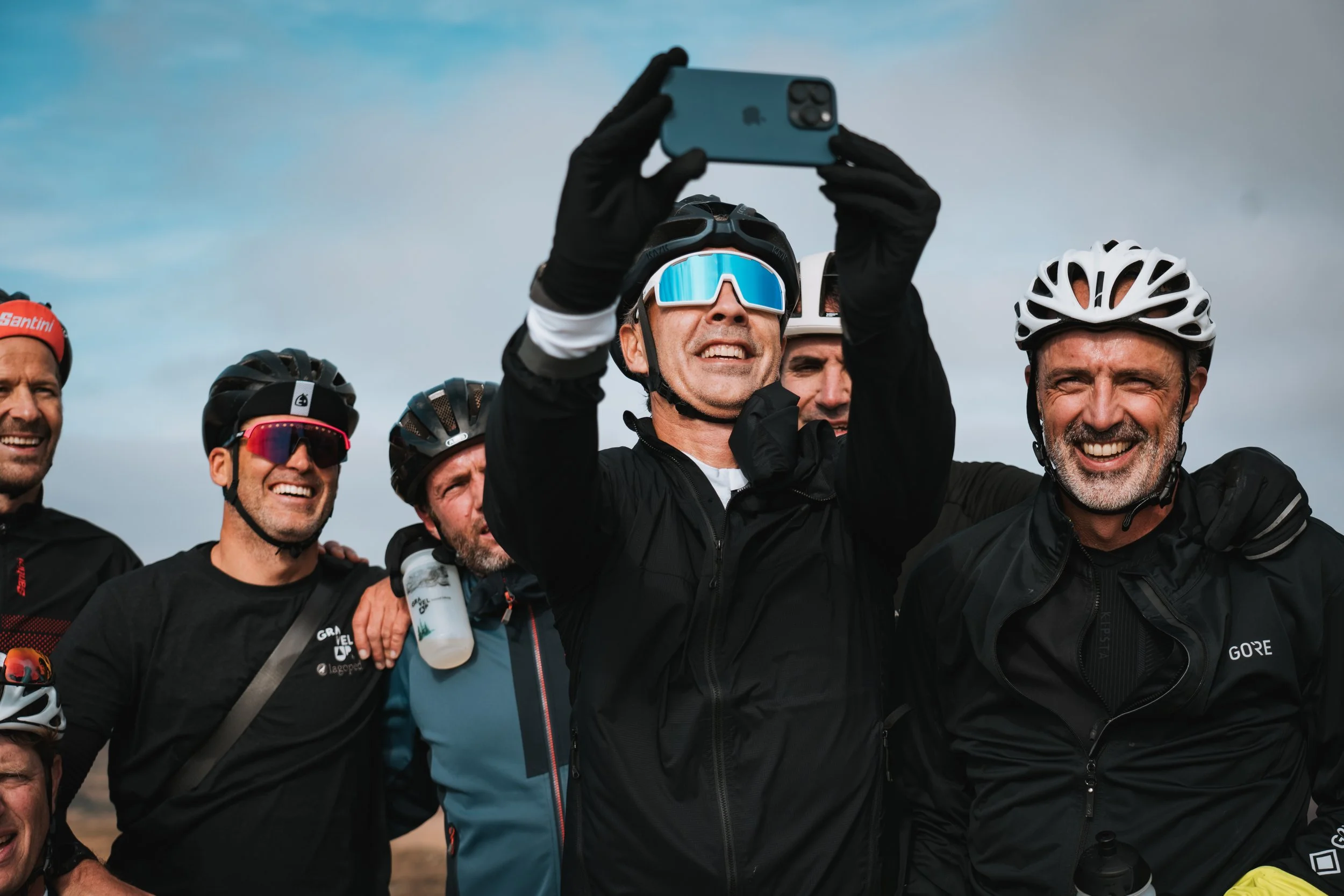 Groupe de cyclistes souriants, portant des casques et des lunettes de soleil, posant pour une photo sous un ciel nuageux.