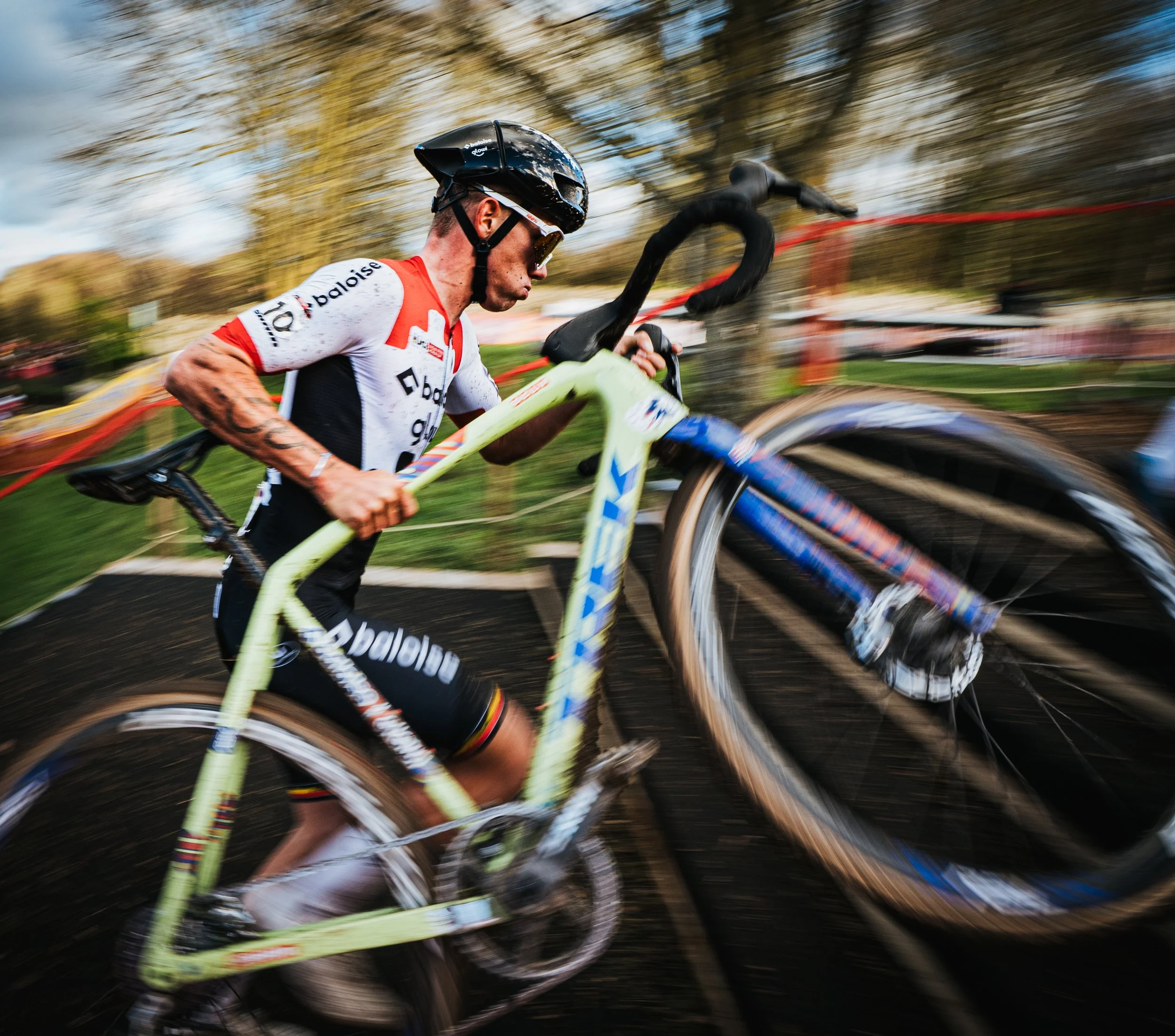 Un coureur cycliste portant un casque noir, une tenue blanche avec le logo 'baloise' et des lunettes de soleil court à grande vitesse lors d'une compétition en plein air, avec un arrière-plan flou montrant des arbres et un ciel nuageux.