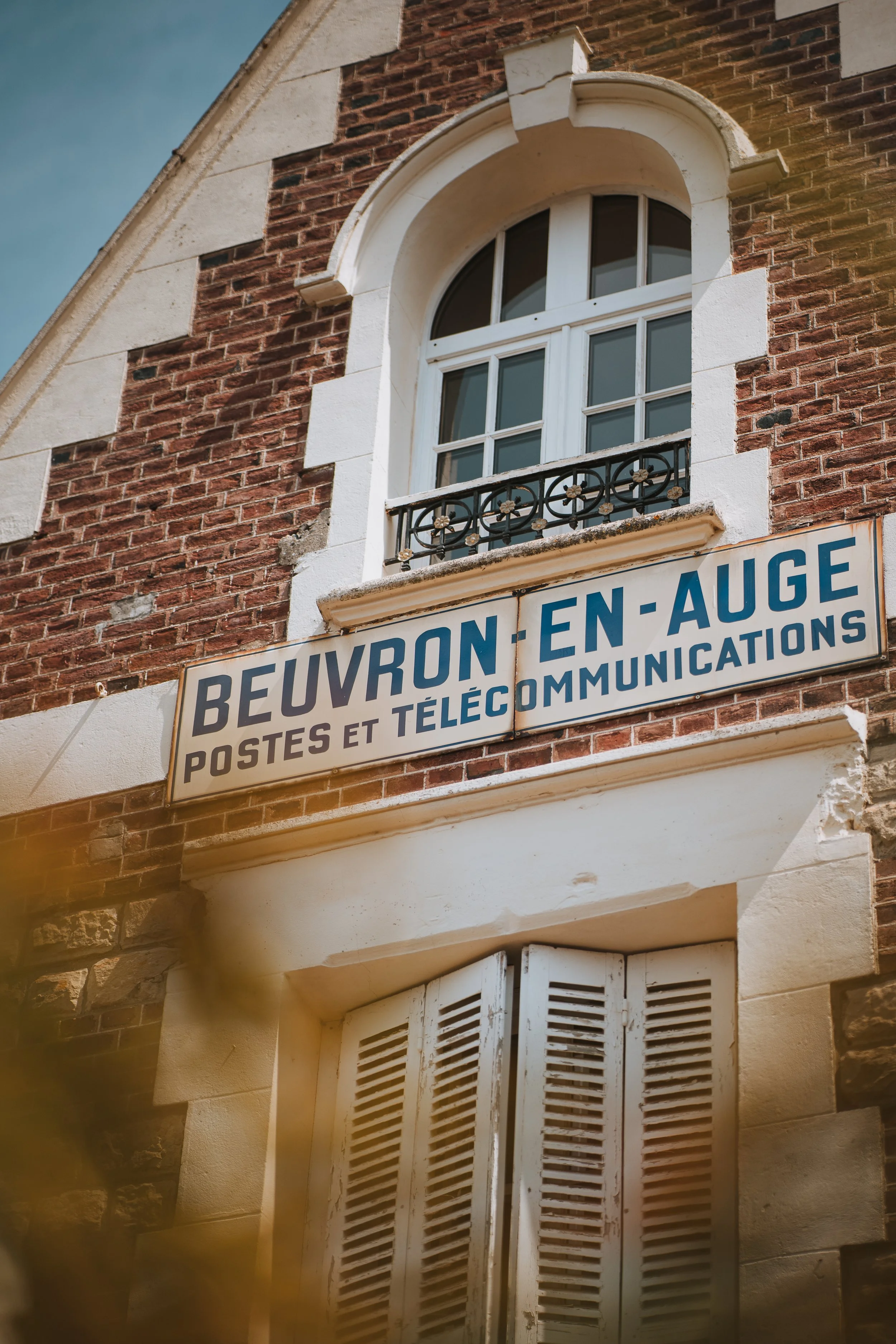 Façade d'un bâtiment en briques avec une fenêtre à volets blancs et un balcon en fer forgé, signalisation commerciale indiquant 'Beuvron-en-Auge, Postes et Télécommunications'.