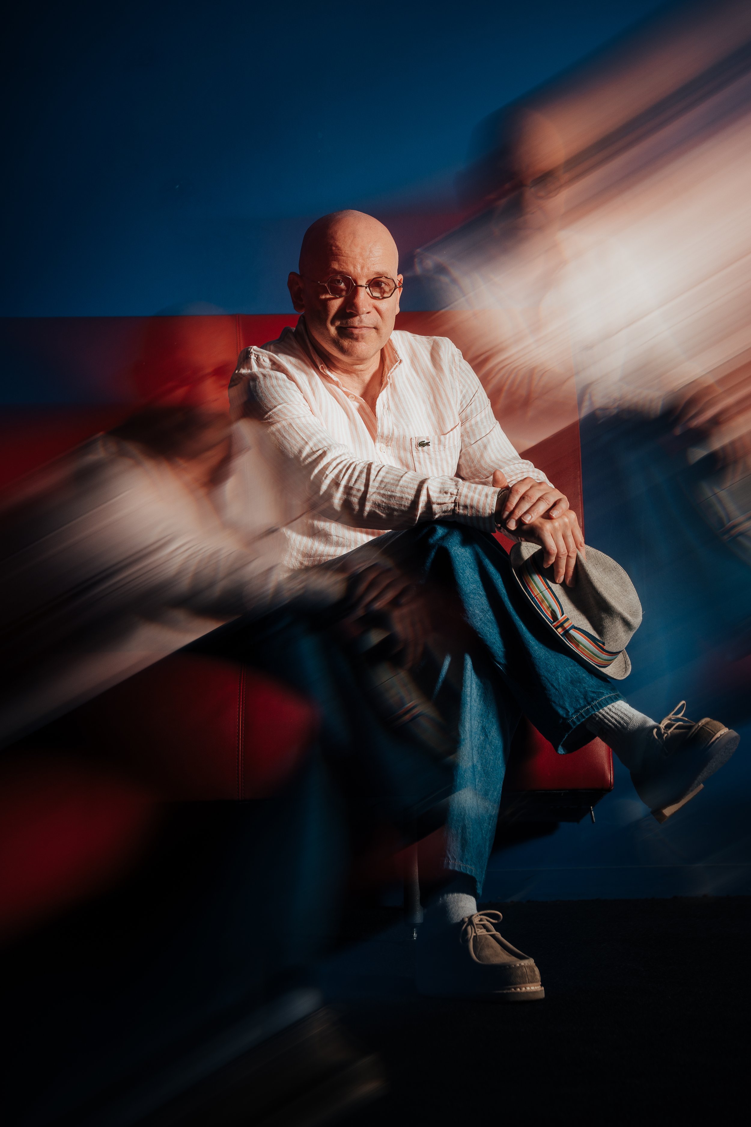 Un homme assis sur une banquette rouge, portant des lunettes, une chemise à rayures et un jean. Il tient un chapeau gris avec un bandeau coloré. La photo semble avoir un effet de mouvement avec des images floues autour de lui.