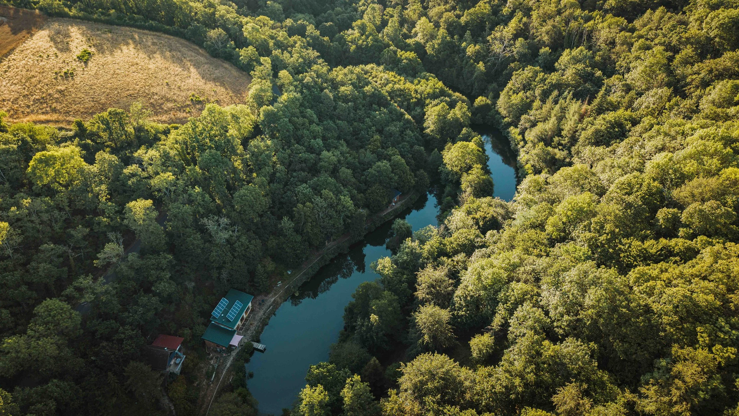 Vue aérienne d'une forêt dense avec une rivière serpentant au milieu, zone en clair avec de la végétation sèche sur le haut gauche, quelques bâtiments dont des panneaux solaires en bas à gauche.