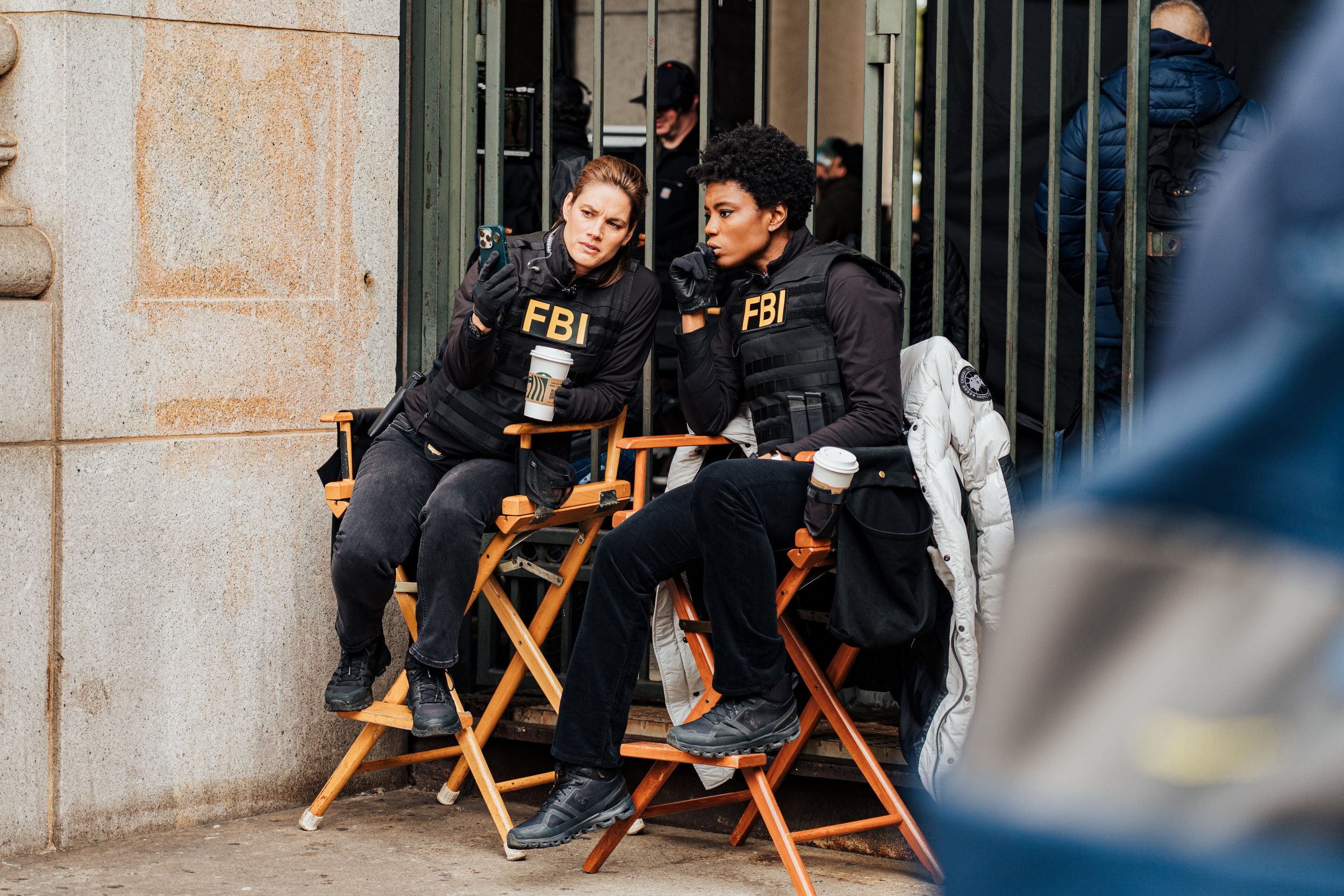 Deux femmes vêtues de vestes FBI discutent et regardent un téléphone tout en assis sur des chaises pliantes, devant un portail en métal avec d’autres personnes en arrière-plan.