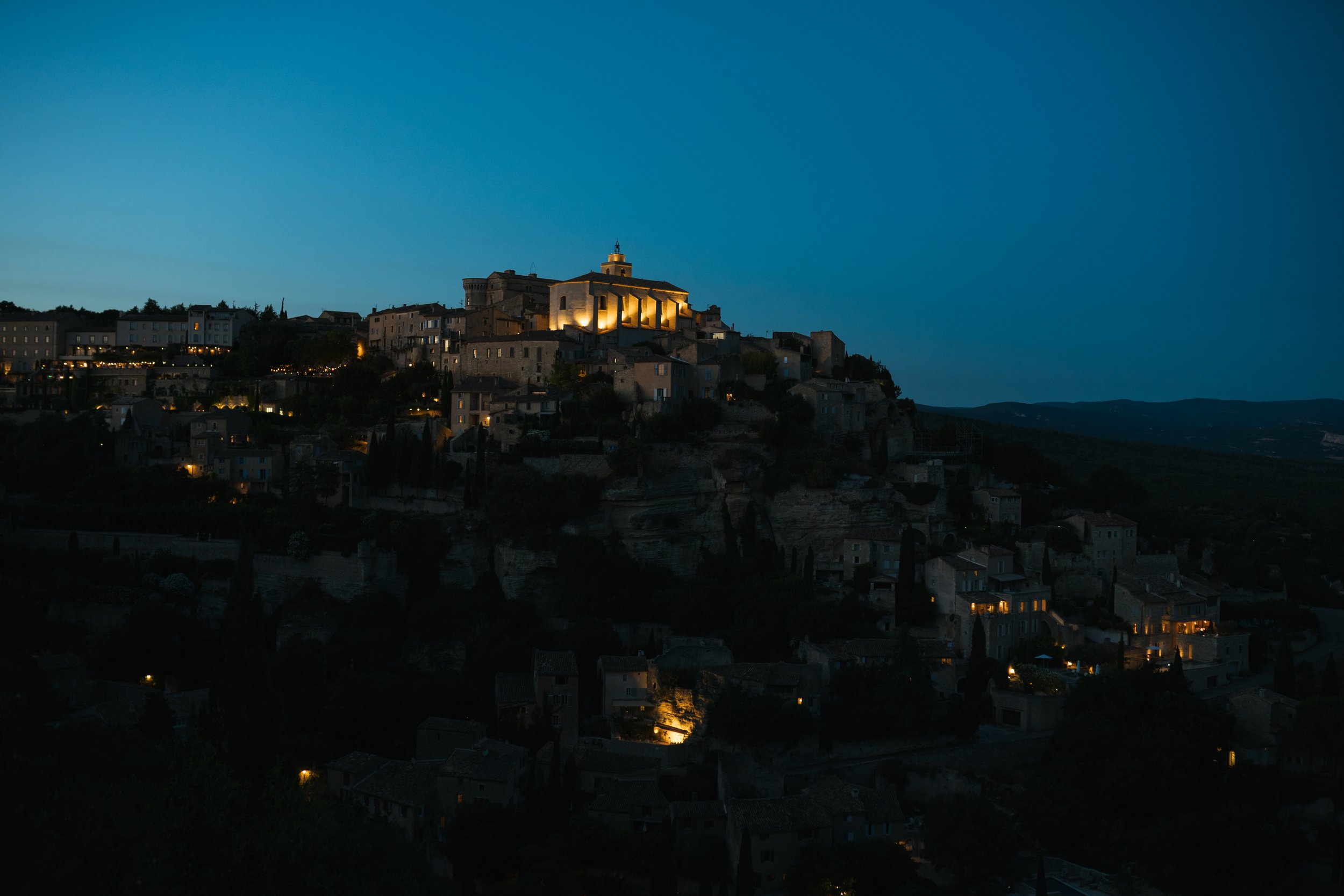 Vue d'une ville perchée sur une colline au crépuscule, avec des bâtiments éclairés par des lumières chaudes et un ciel bleu foncé.