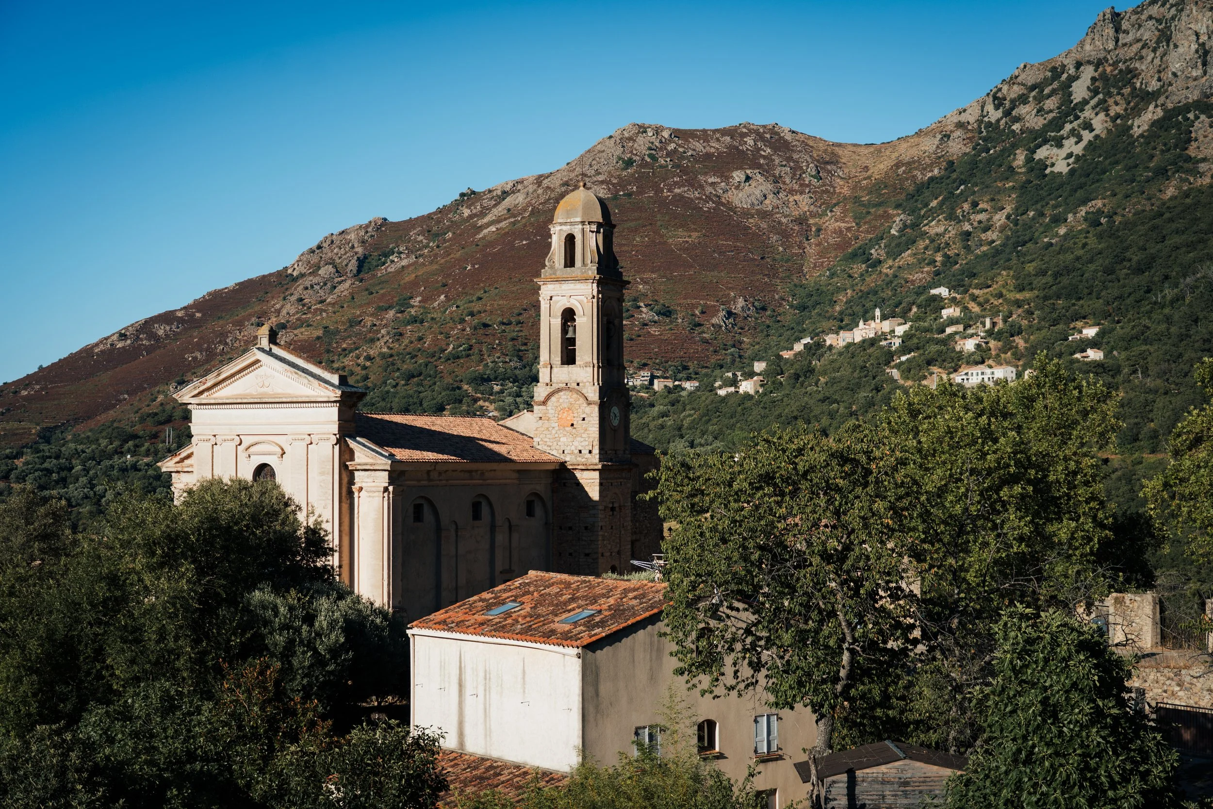 Une église ancienne avec une tour de clocher en pierre, située dans un village au pied de montagnes avec des maisons dispersées sur la colline. La scène est en plein jour avec un ciel clair.