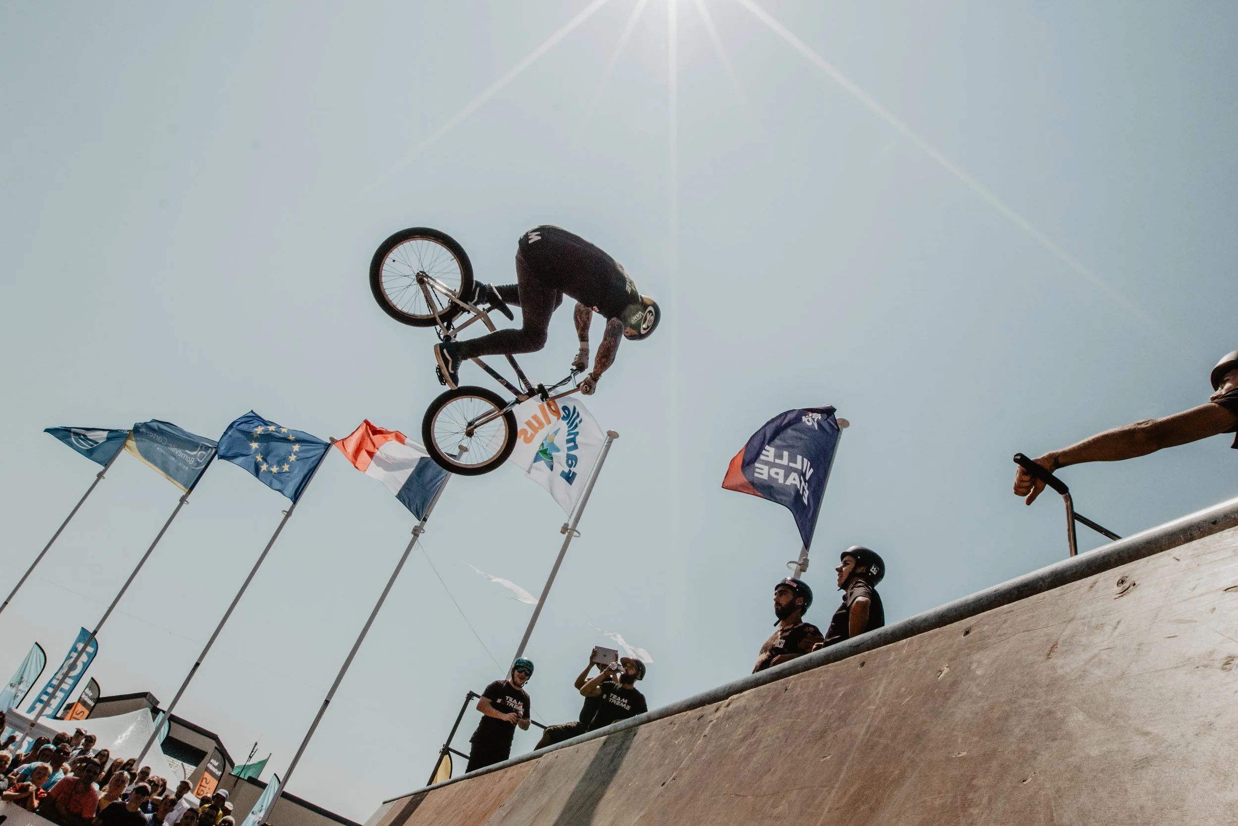 Un rider effectuant une figure de BMX en l'air lors d'une compétition, entouré de spectateurs et de drapeaux. Le ciel est clair et ensoleillé.