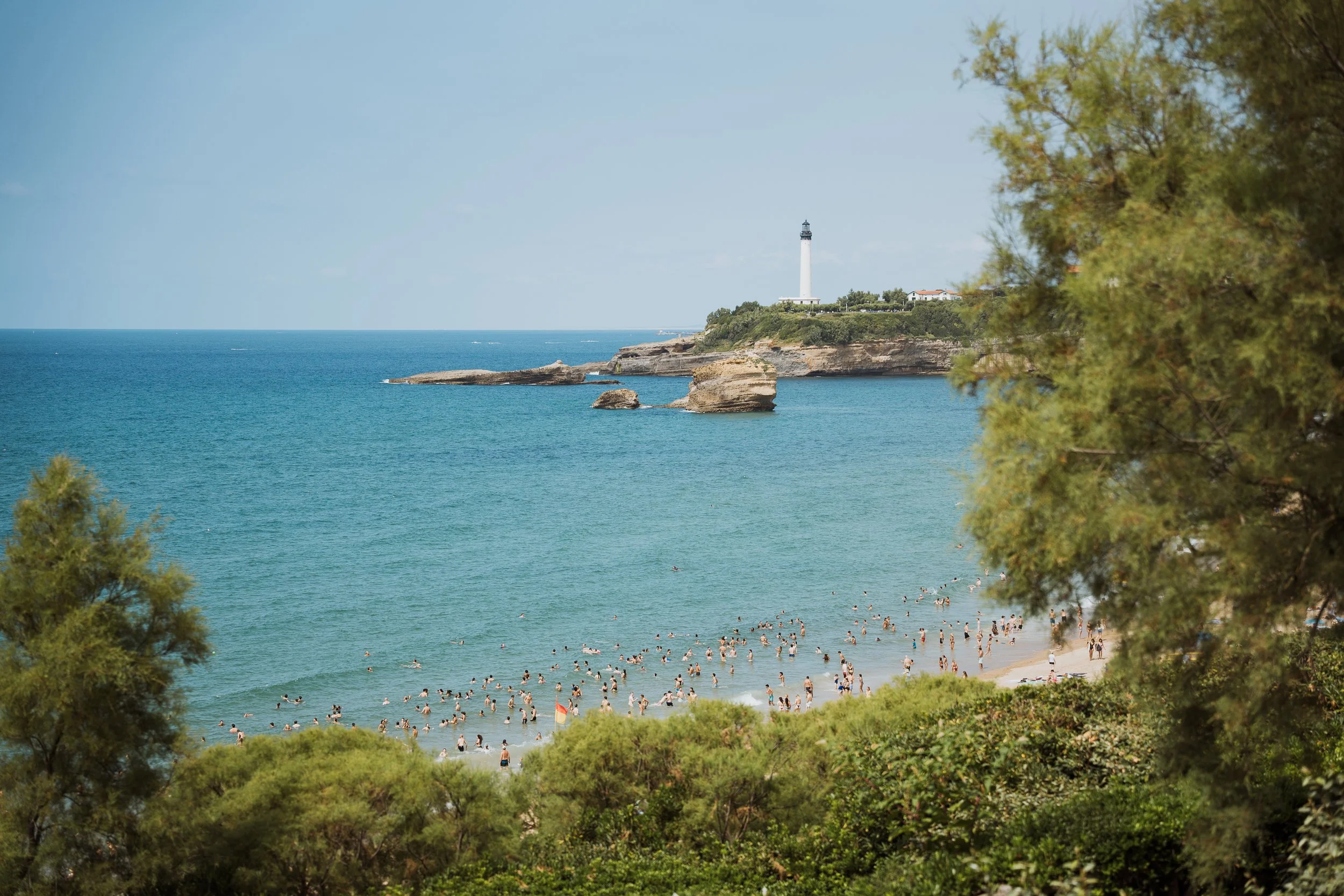 Plage bondée avec des personnes nageant dans la mer, à proximité d'une falaise avec un phare blanc et une maison en arrière-plan, entourée d'arbres verts.