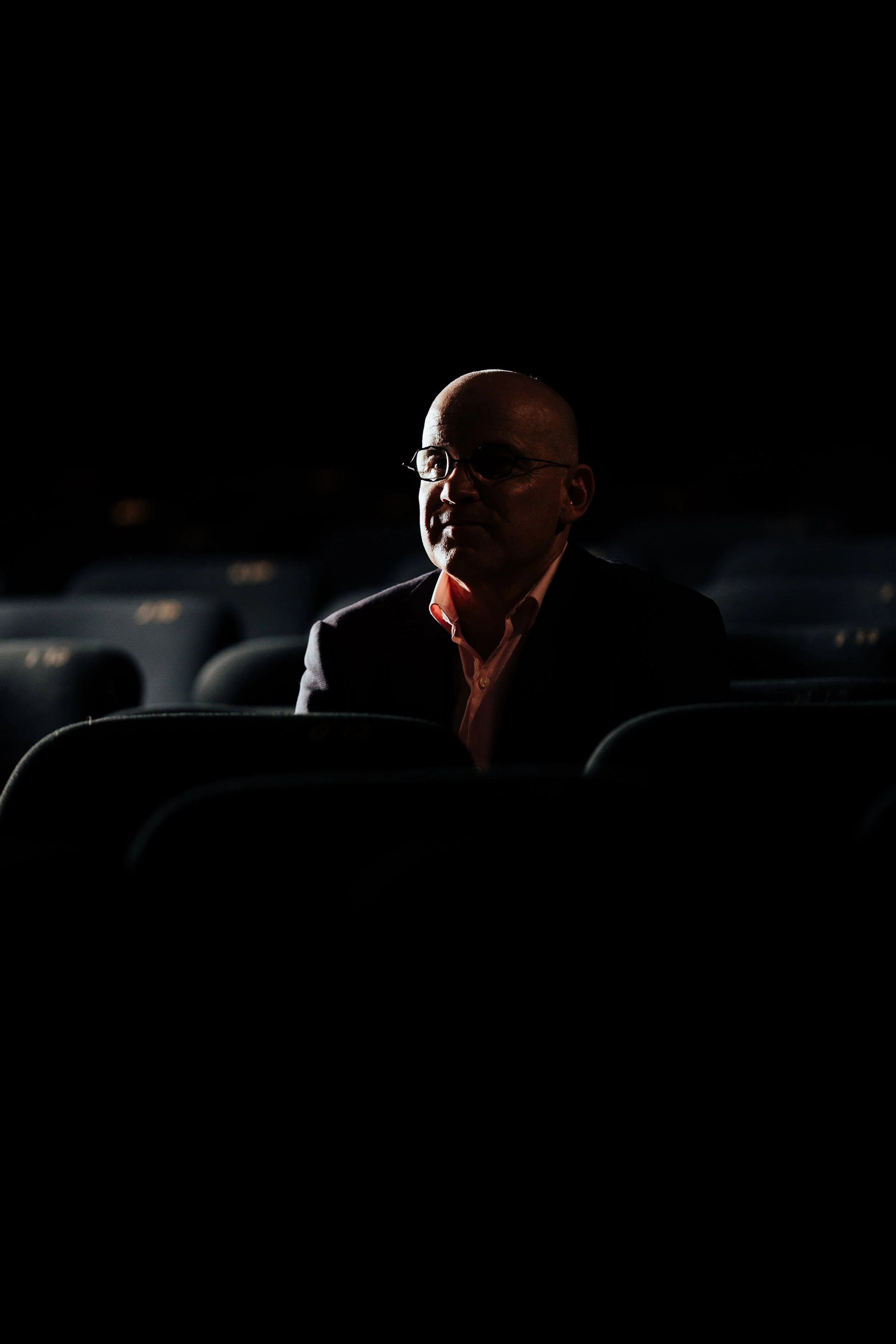 Un homme portant des lunettes, assis dans une salle sombre, éclairé partiellement par une lumière venant de l'avant.
