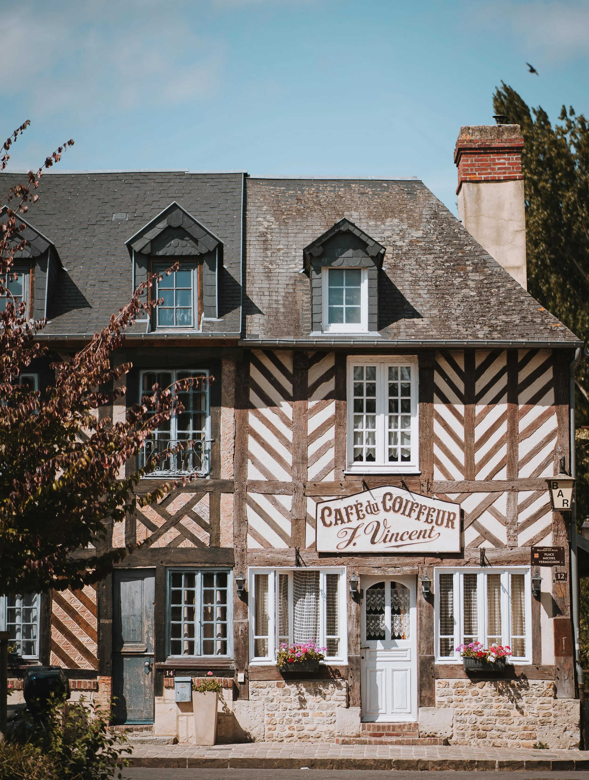 Façade d'une maison ancienne en style traditionnel, avec fenêtres à petits carreaux, volets en bois, fleurs dans des jardinières et enseigne indiquant un café nommé 'Café du Coiffeur F. Vincent'.