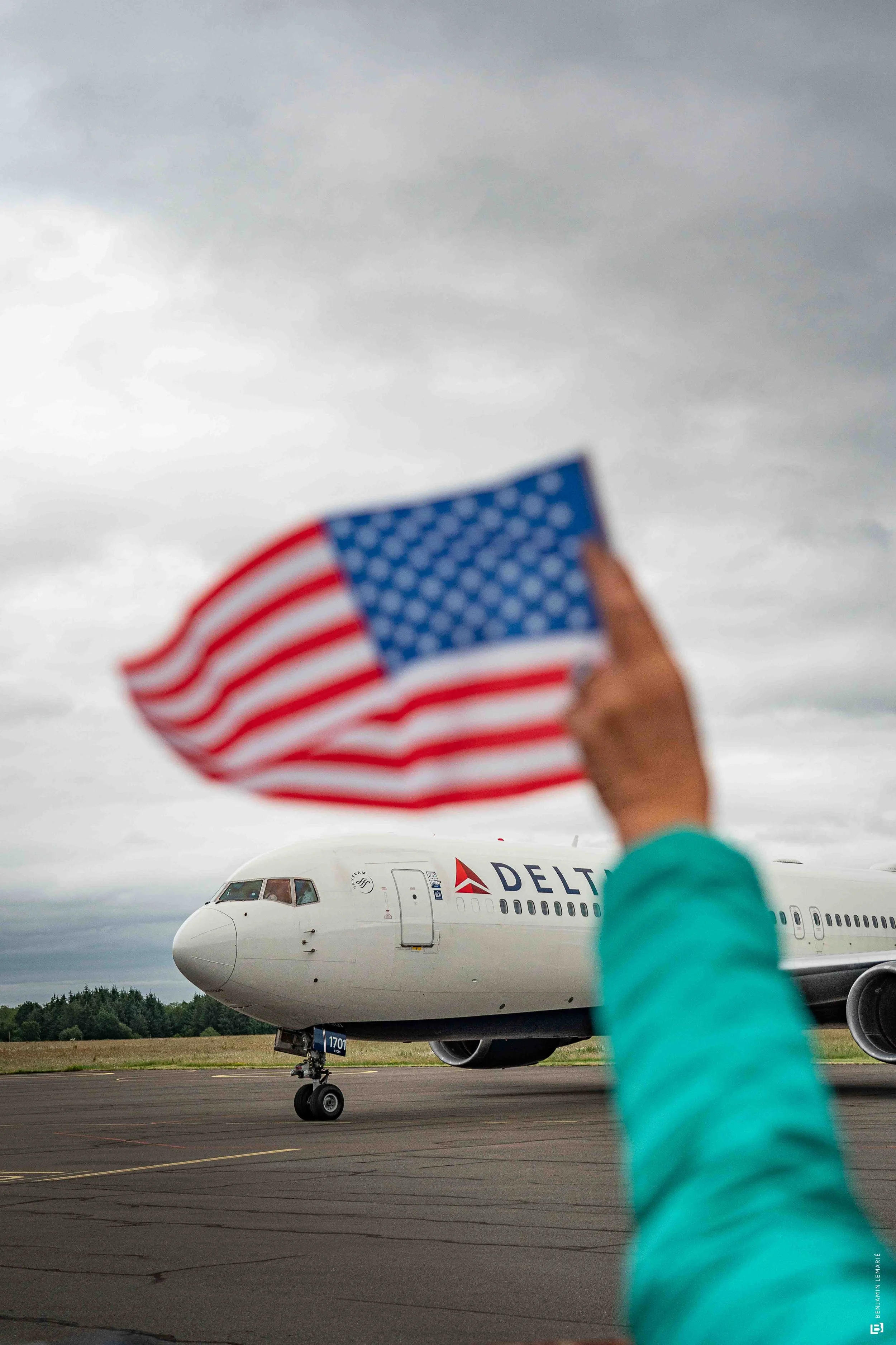 Un avion au sol avec le logo Delta visible, une personne en arrière-plan agite un drapeau américain, le ciel est nuageux.