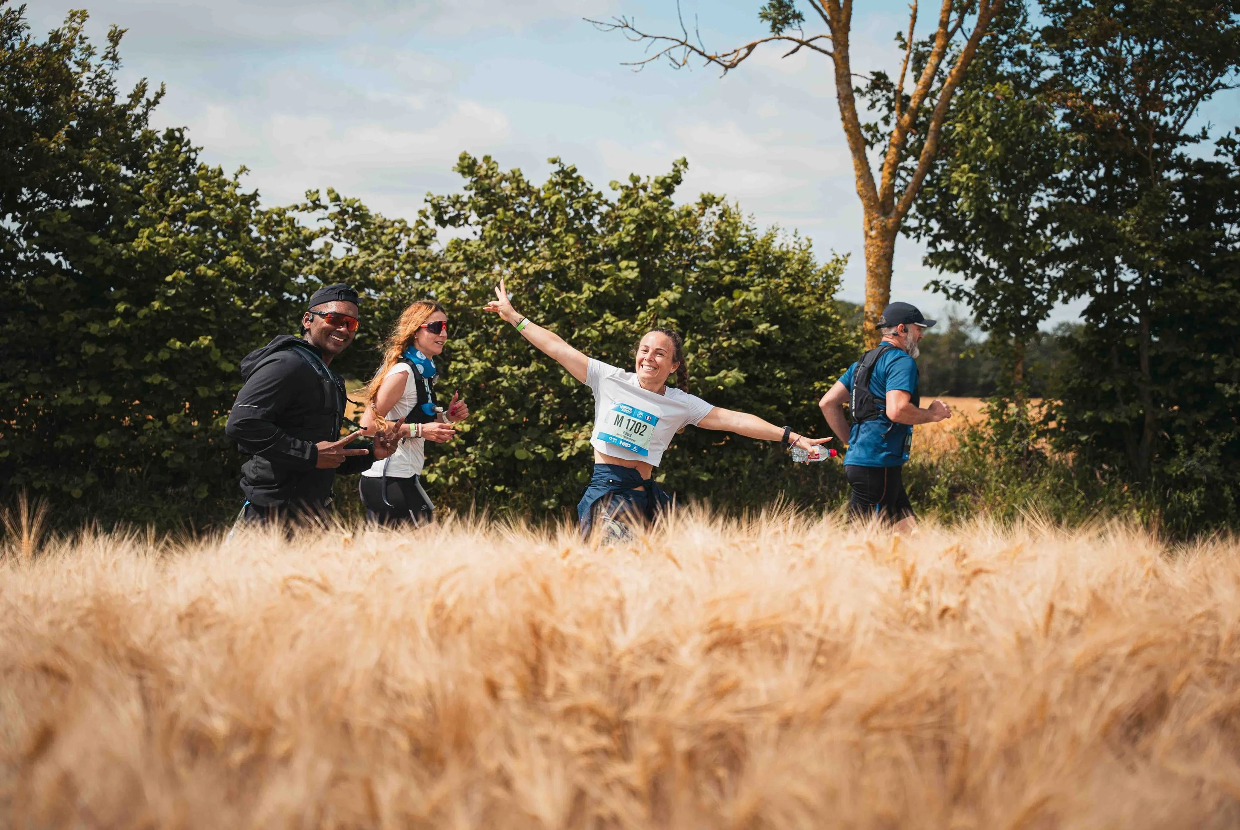 Groupe de quatre personnes en train de courir ou marcher en plein air dans un champ, souriants et portant des vêtements de sport, avec des arbres en arrière-plan.