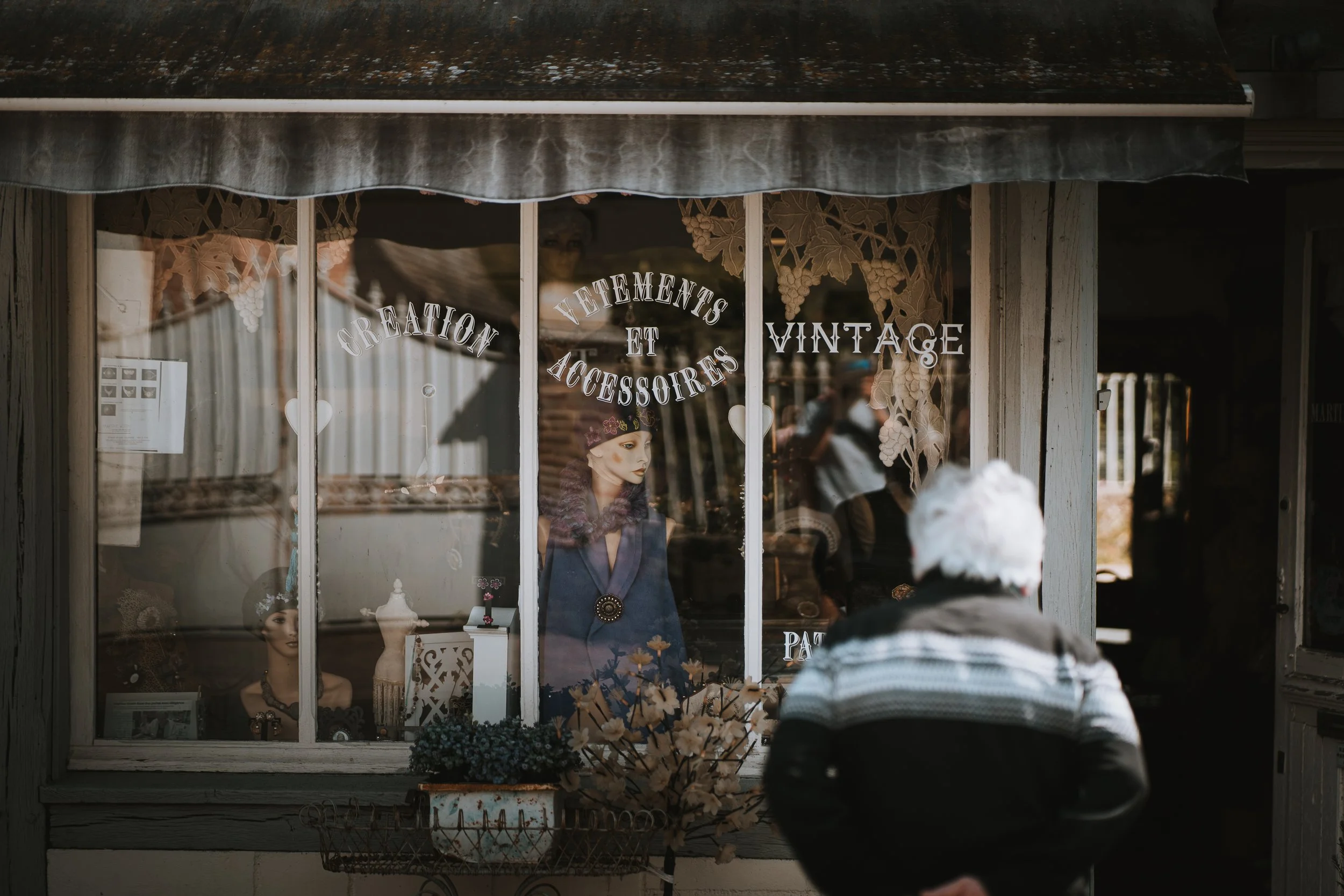 Une vitrine d'une boutique vintage avec des mannequins et des plantes en décoration, affichant des accessoires et vêtements anciens.