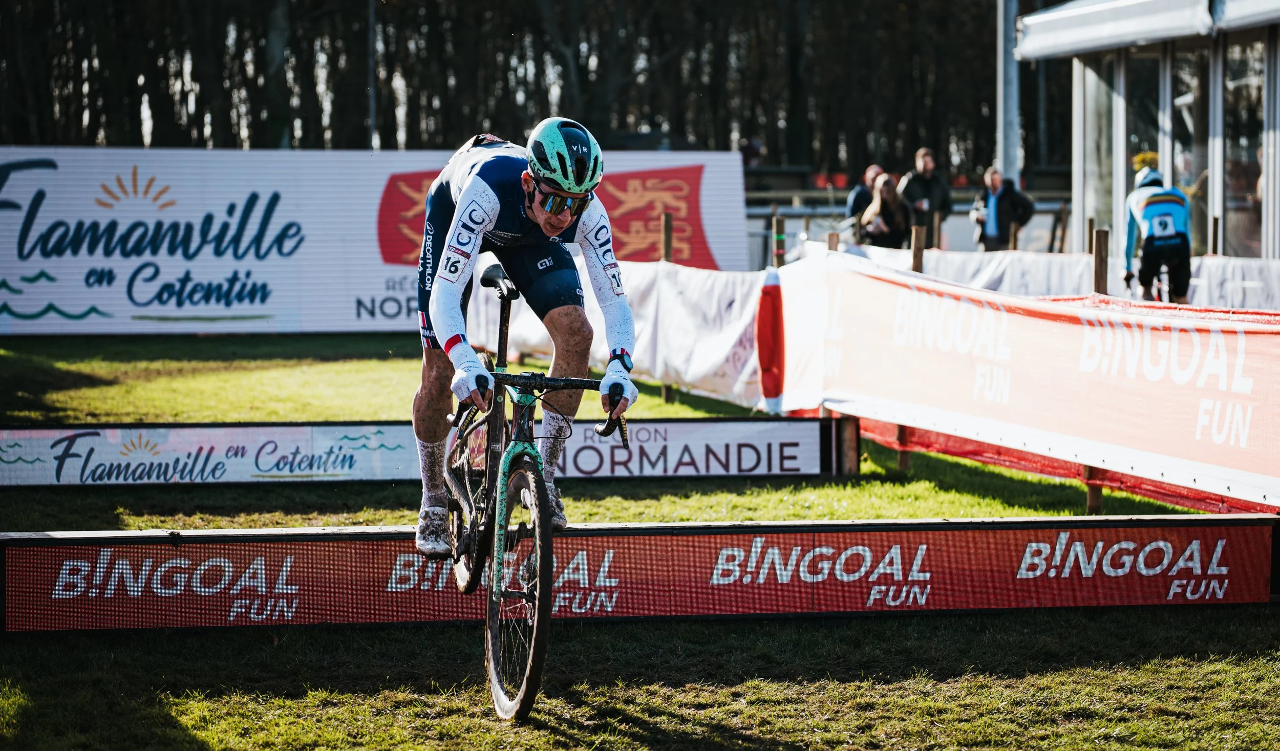 Coureur de vélo cycliste en plein effort lors d'une compétition, portant un casque bleu et des vêtements de course, traversant une ligne d'arrivée ou de passage sur un parcours en extérieur avec des spectateurs en arrière-plan.