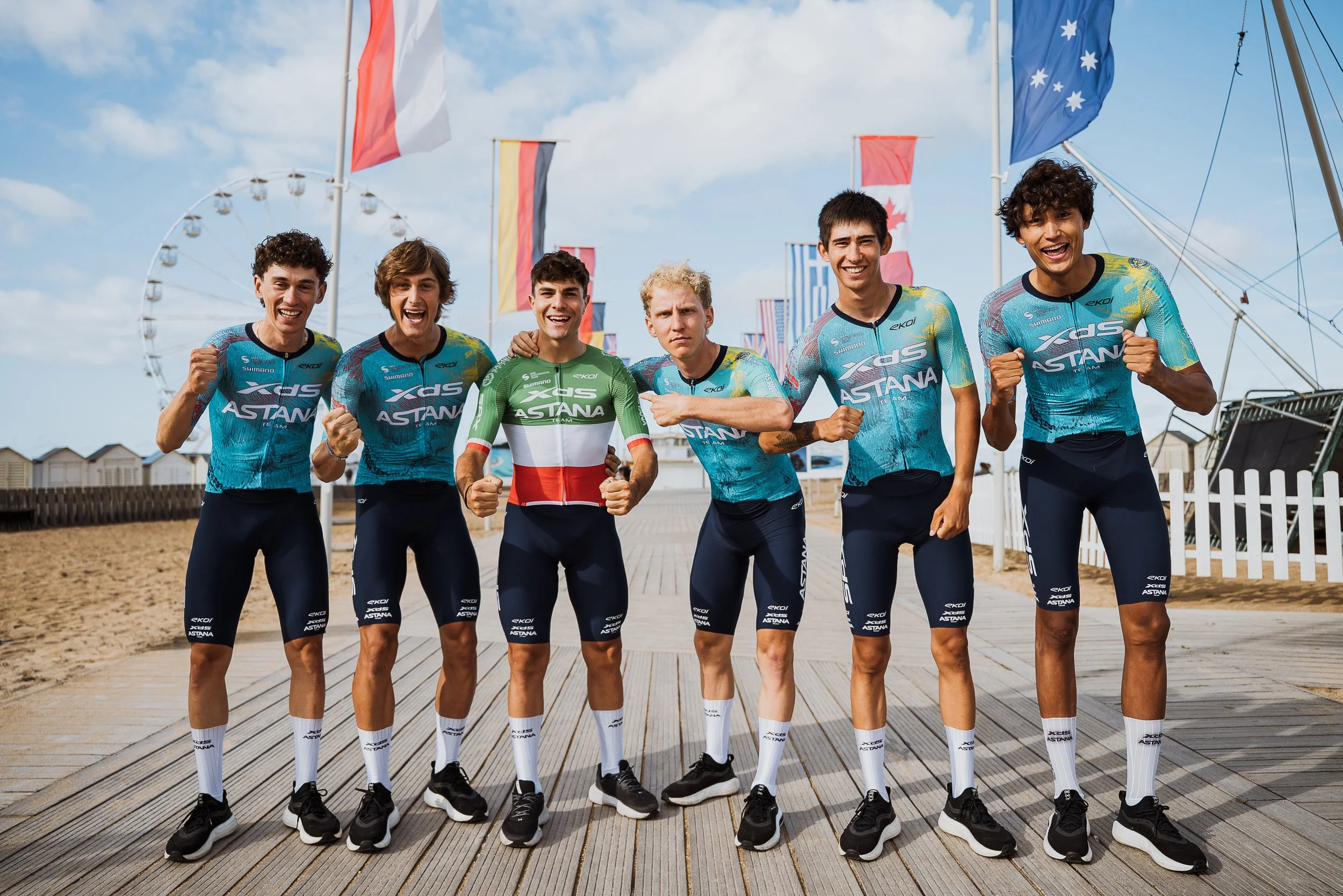 Un groupe de sept jeunes athlètes en tenues de sport de l'équipe Astana pose en souriant sur un pont en bois, avec des drapeaux colorés en arrière-plan et un grand rouleau de ferris en forme de roue dans le fond, par un temps ensoleillé avec des nuages