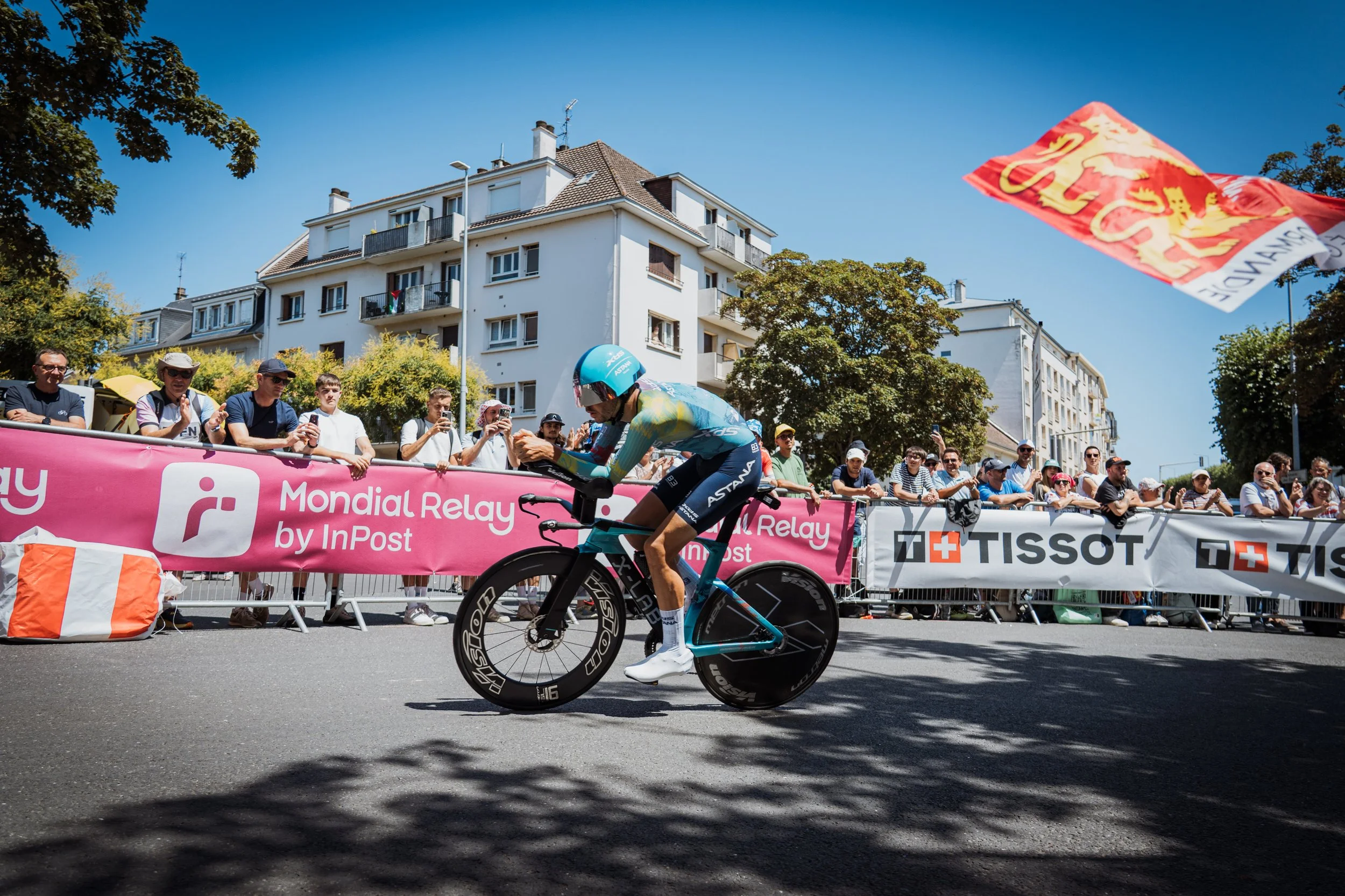 Un cycliste professionnel en pleine course lors d'une compétition, avec un groupe de spectateurs derrière une barrière, en plein jour avec un ciel clair et un immeuble résidentiel en arrière-plan.