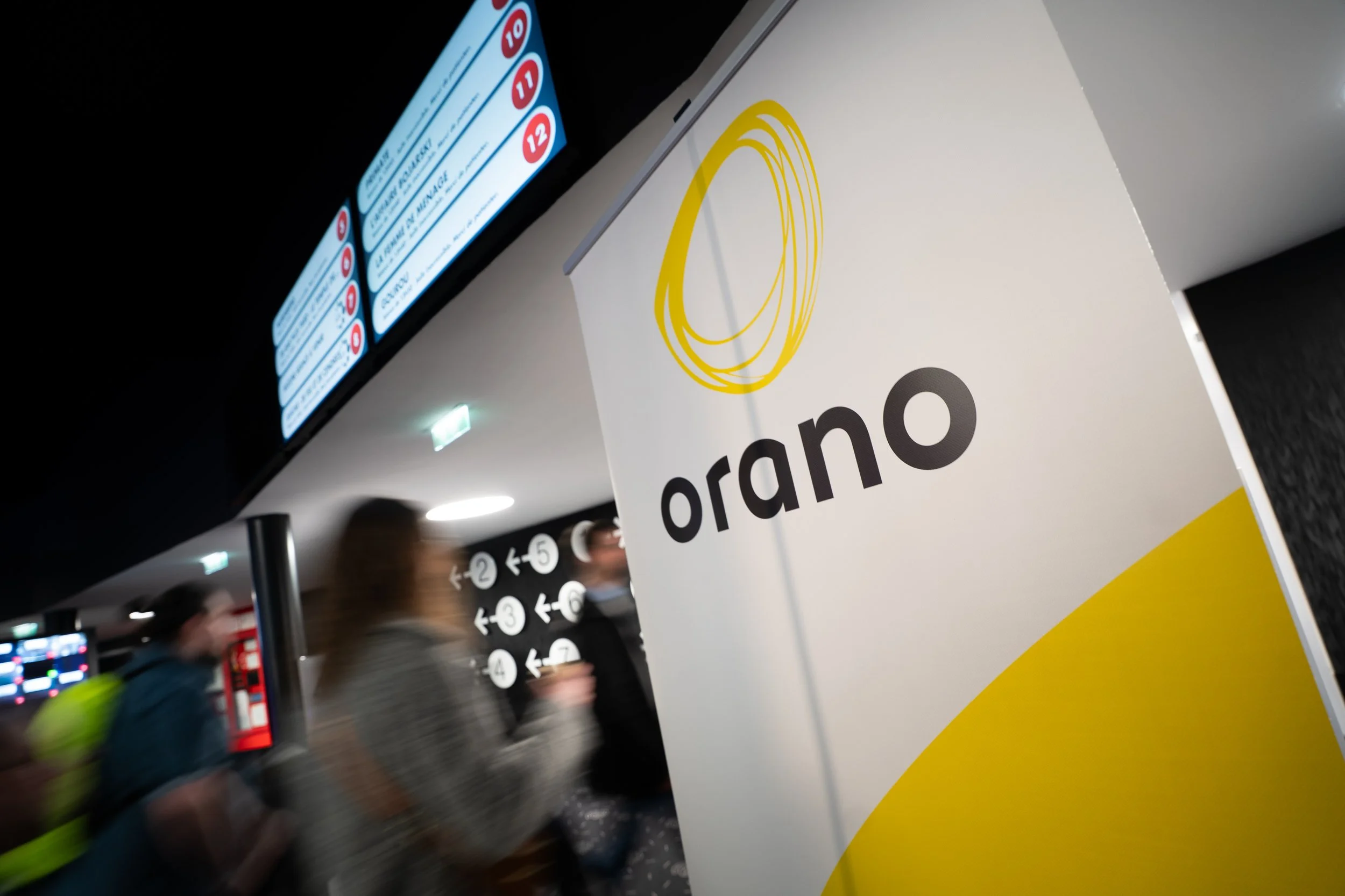 Vue partielle d'une salle d'aéroport avec un panneau blanc portant un logo jaune et le mot 'Grand' en noir, des écrans d'information en haut avec des chiffres rouges et blancs, et des passagers flous en mouvement.